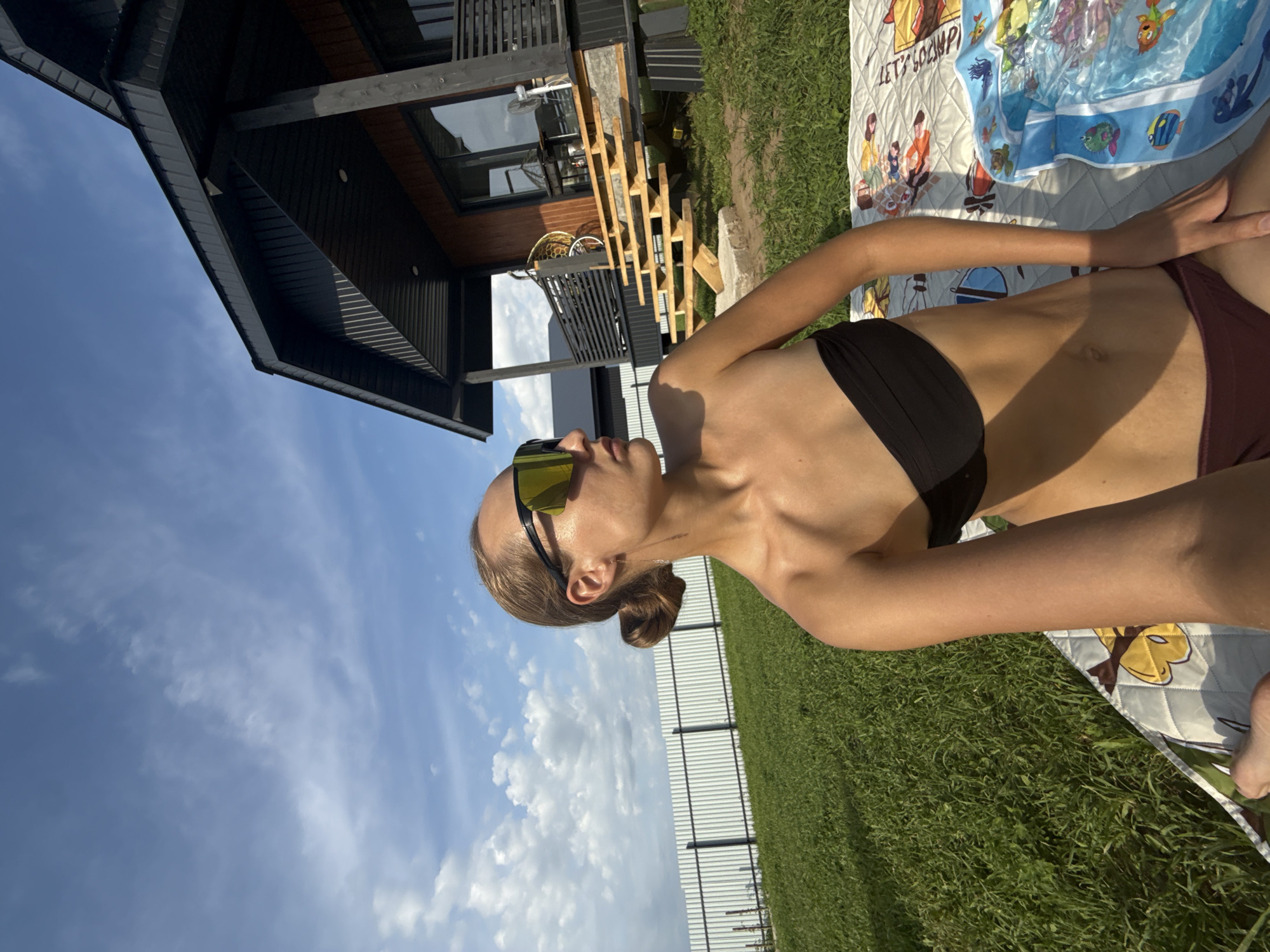 In the city of Podshivalovo, Russia, a woman is pictured on a sunny day in a grassy yard. She is seated on a blanket next to an inflatable water toy, wearing a black bandeau bikini top and burgundy bikini bottoms. She has on large, reflective sunglasses and her blonde hair is pulled back in a bun. Behind her, a modern wooden house with a dark roof and exposed wooden stairs stands under a bright blue sky with scattered clouds. A white metal fence runs along the edge of the property.