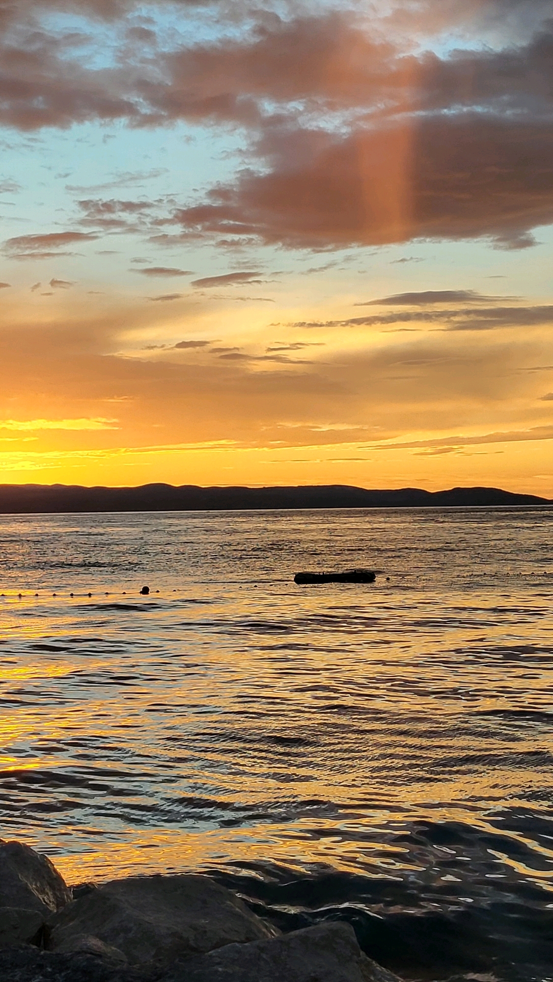 The image depicts a serene sunset over the Adriatic Sea, viewed from the shoreline of Općina Tučepi, Croatia. The sky is painted with vibrant hues of orange and yellow, transitioning into soft blues and grays towards the top. Wispy clouds catch the fading sunlight, with a notable vertical shaft of light piercing through a cluster of darker clouds. In the distance, the silhouette of a mountainous coastline is visible against the horizon. The water's surface is rippled, reflecting the warm tones of the sky, creating a shimmering effect. A long, dark object, possibly a floating platform or debris, is present on the water. A series of buoys or floats are strung together in a line, also on the water's surface, closer to the shore. The foreground features large, dark rocks, partially submerged at the water's edge, suggesting a natural, undeveloped coastline. The overall scene is tranquil and evokes a sense of calm.