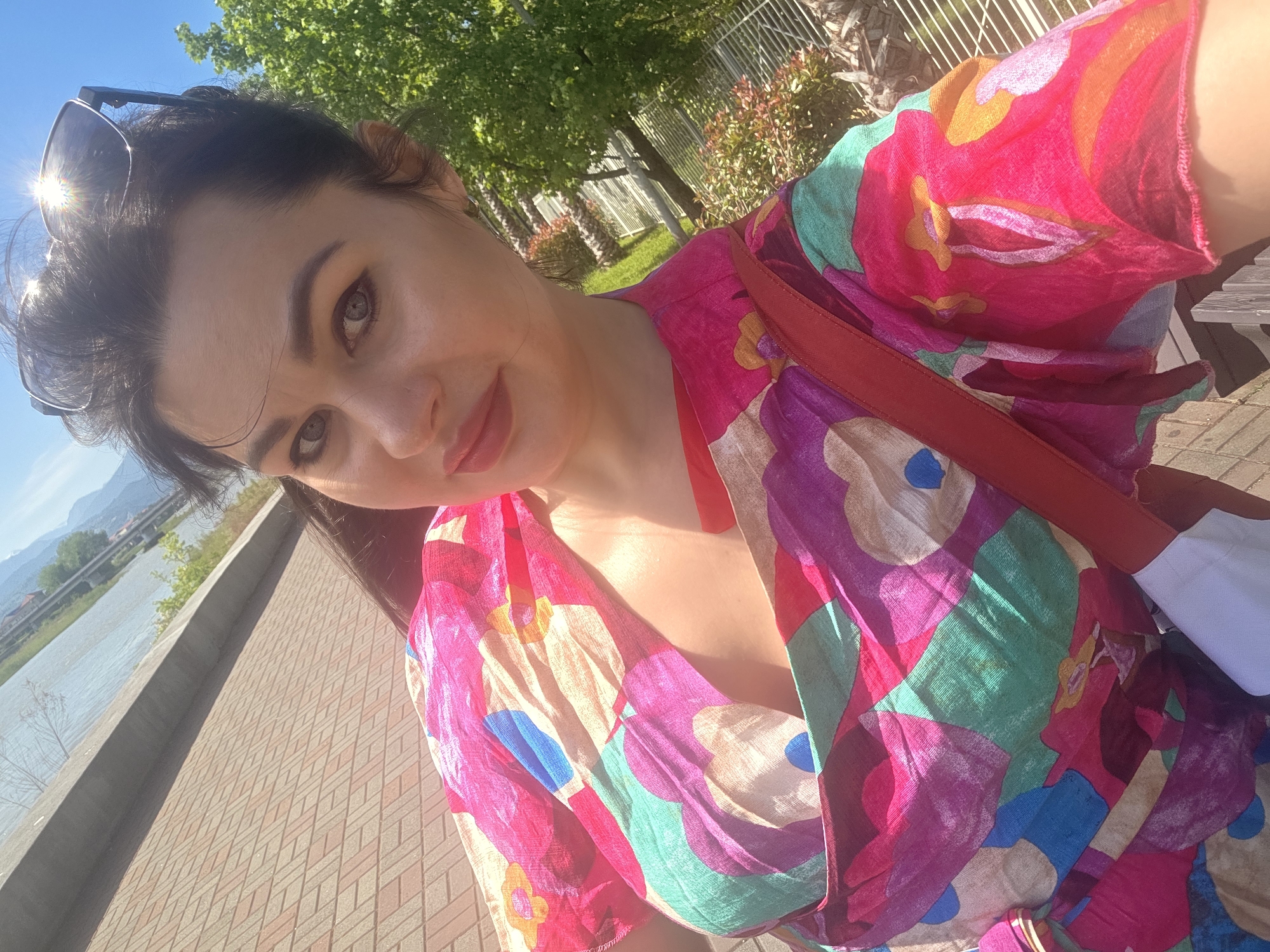 An adult female is depicted in a close-up, outdoor selfie taken on a clear, sunny day in what appears to be the city of Sochi, Russia. The woman, positioned centrally, has dark hair pulled back, light grey-blue eyes, defined dark eyebrows, and wears light reddish-pink lipstick. Dark-rimmed sunglasses are pushed up onto her head, reflecting a prominent sunburst. She is wearing a vibrant, loose-fitting garment with an abstract pattern composed of fuchsia, magenta, teal, purple, blue, and orange shapes. A red strap, presumably from a bag, crosses over her right shoulder, and a small white object is partially visible below it.
The immediate foreground features reddish-brown brick herringbone pavement. Behind the woman, a low concrete barrier separates the paved area from a wide body of water, likely the Black Sea or a river flowing into it, reflecting the bright blue sky. On the far side of the water, green mountains and indistinct urban structures are visible. To the right of the woman, dense green foliage, including tall trees and bushes, is brightly illuminated by direct sunlight, creating strong highlights and shadows. A white metal fence or railing is partially visible on the far right. The overall setting, with its mountainous backdrop, large body of water, and subtropical vegetation, is consistent with the coastal city of Sochi, Russia.