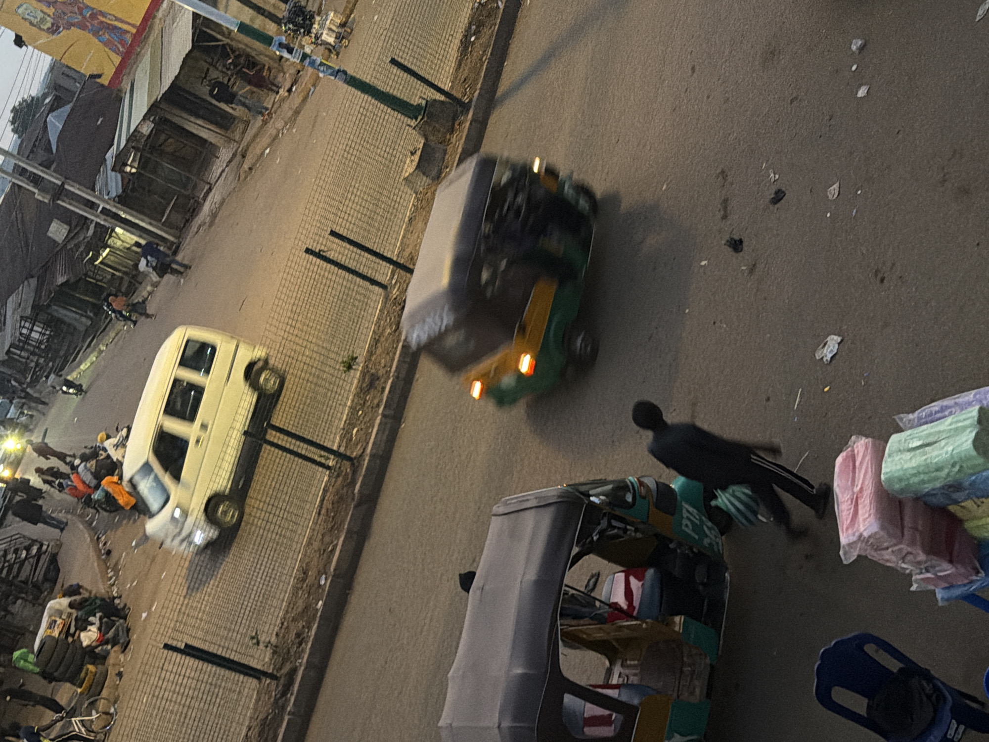 The image depicts a street scene in Bukuru, Nigeria, with a focus on vehicular and pedestrian activity. In the foreground, a grey and green auto-rickshaw is parked, with a person in dark clothing walking away from it, carrying a striped green bag. Next to the auto-rickshaw, a stack of colorful wrapped goods, possibly textiles, is visible, along with a blue plastic chair.
Further down the street, a white minibus is in motion, appearing to be a public transport vehicle. Several people are seen on the side of the road, near stalls or shops with corrugated iron roofs. Other auto-rickshaws and motorcycles are also present, indicating a busy thoroughfare. The environment appears to be an urban setting with a mix of informal market stalls and more permanent structures. The lighting suggests it is either early morning or late afternoon, with a muted, natural light. The ground is paved, and a metal fence runs along a portion of the street. Debris is scattered on the road and sidewalk.