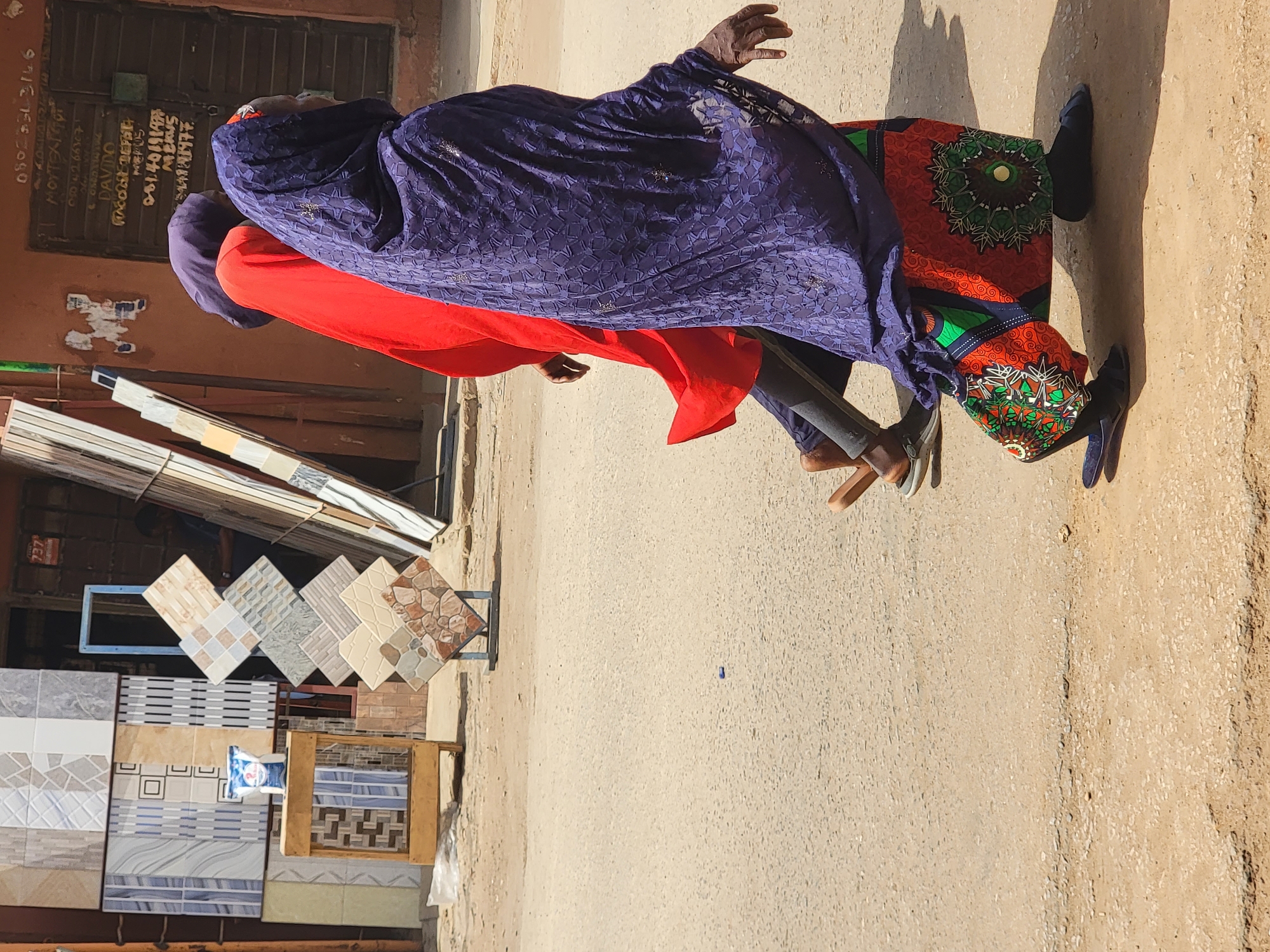 Two individuals walk past a store displaying ceramic tiles in Maiburiji, Nigeria. One person wears a red garment with a blue lace shawl. The other wears a patterned orange skirt. The street is unpaved. The store has its business information written on the wall.