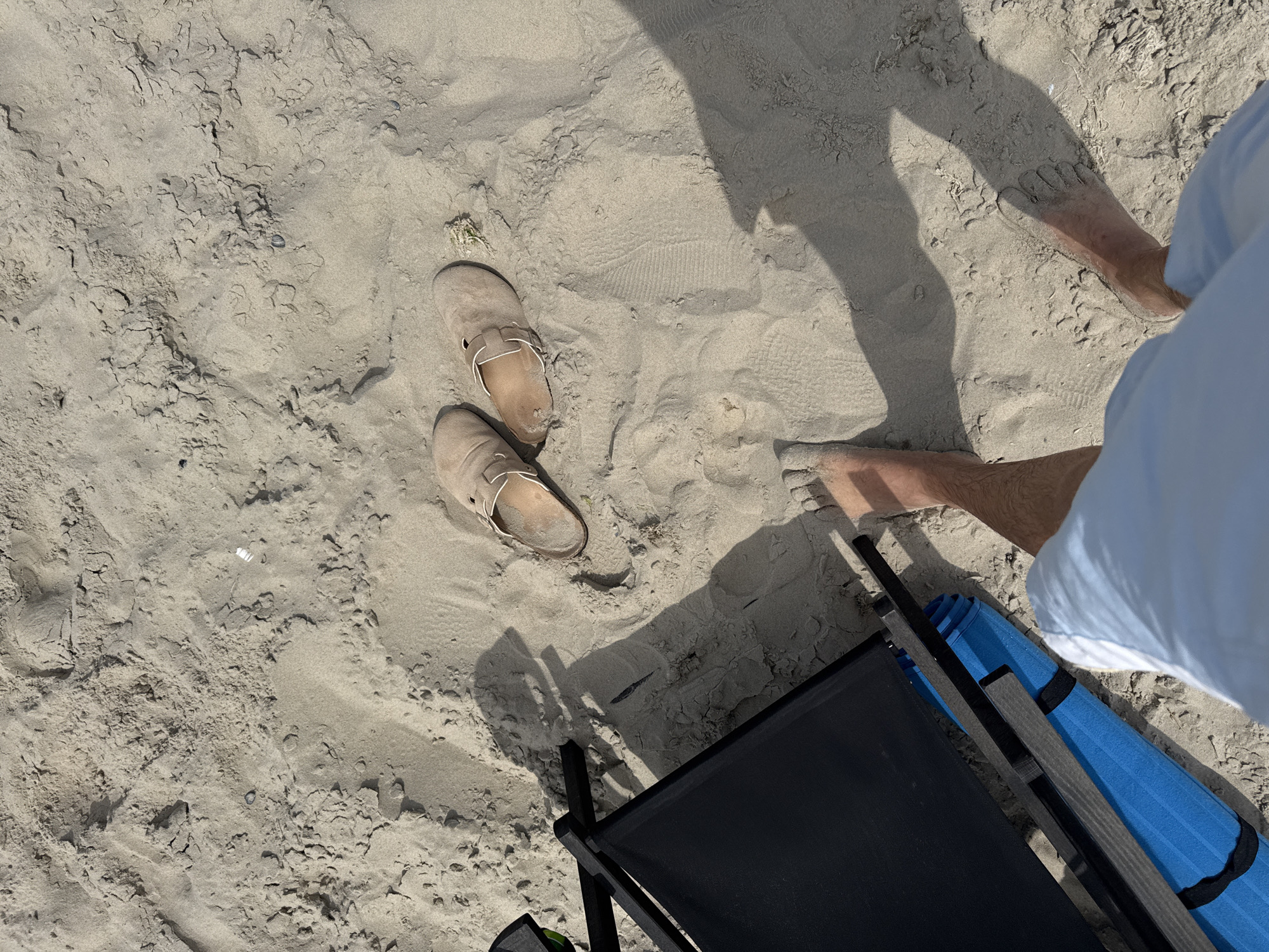 Two tan slip-on shoes rest on a sandy surface, positioned next to a black beach chair and a blue yoga mat. The sand is uneven and textured, with visible footprints and debris. A person's legs and feet are visible in the foreground, suggesting they are standing or walking on the sand. The scene appears to be outdoors, possibly on a beach given the sand and beach chair. The image is shot from a high angle, looking down at the scene. The lighting suggests it is daytime, with shadows cast by the beach chair and the person. The overall impression is one of a relaxed, outdoor setting. This image was taken on the beach in Langeoog, Germany.
