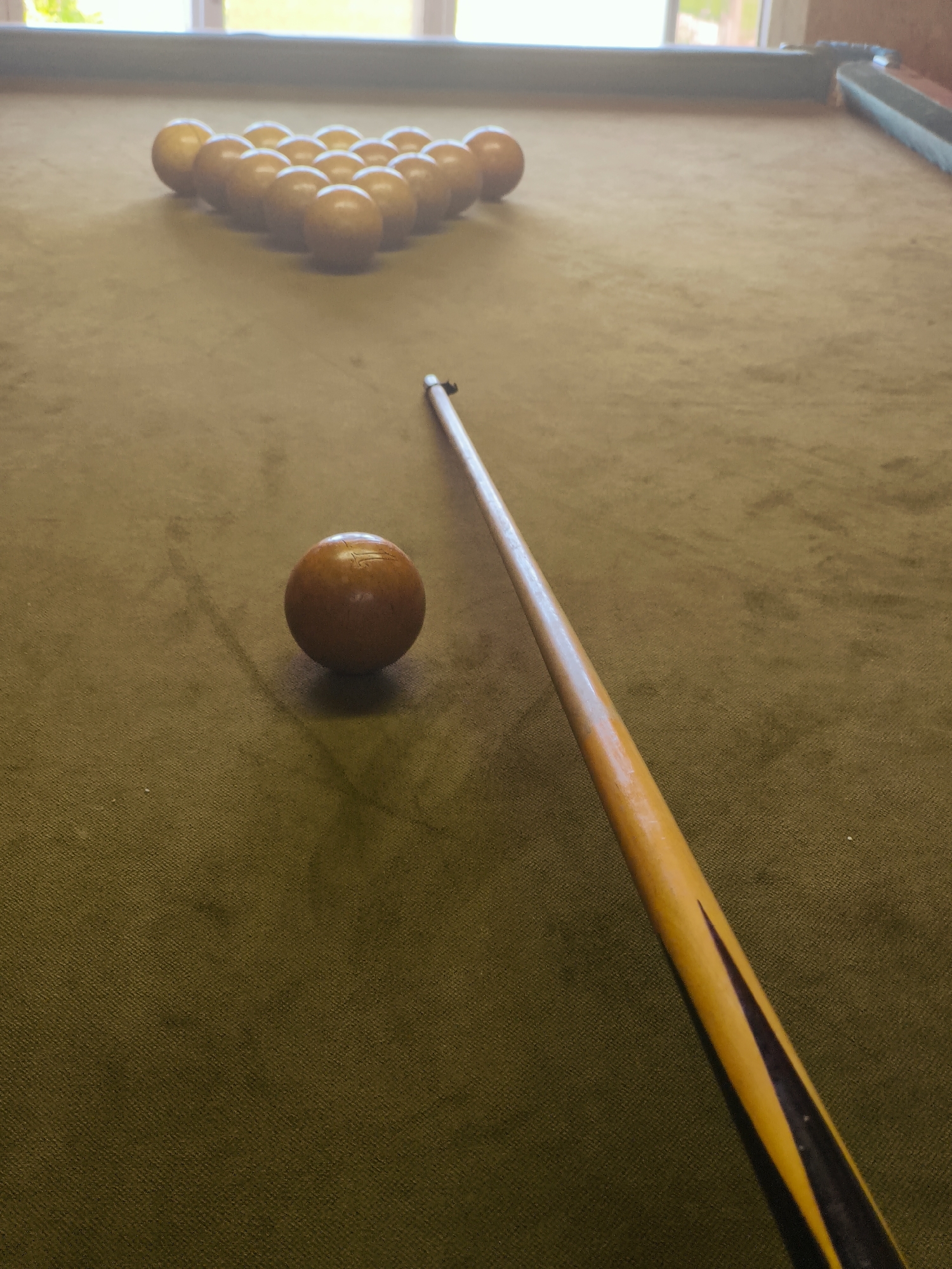 The image presents a high-angle view of a pool table situated indoors in Терновский округ, Ukraine. The table is covered in a textured, olive-green felt. Fifteen solid, light-brown pool balls are arranged in a triangular rack formation at the far end of the table. A single, larger, light-brown cue ball is positioned in the foreground, offset to the left of the central axis. A pool cue, predominantly yellow with dark triangular inlays along its lower shaft, extends diagonally across the felt from the bottom right to the middle left, its tip pointing towards the cue ball. The dark-colored rails of the pool table are visible along the upper perimeter of the frame. In the background, a window with white mullions is present, admitting diffused natural light into the room and revealing blurred outdoor greenery. No individuals are visible in the scene.