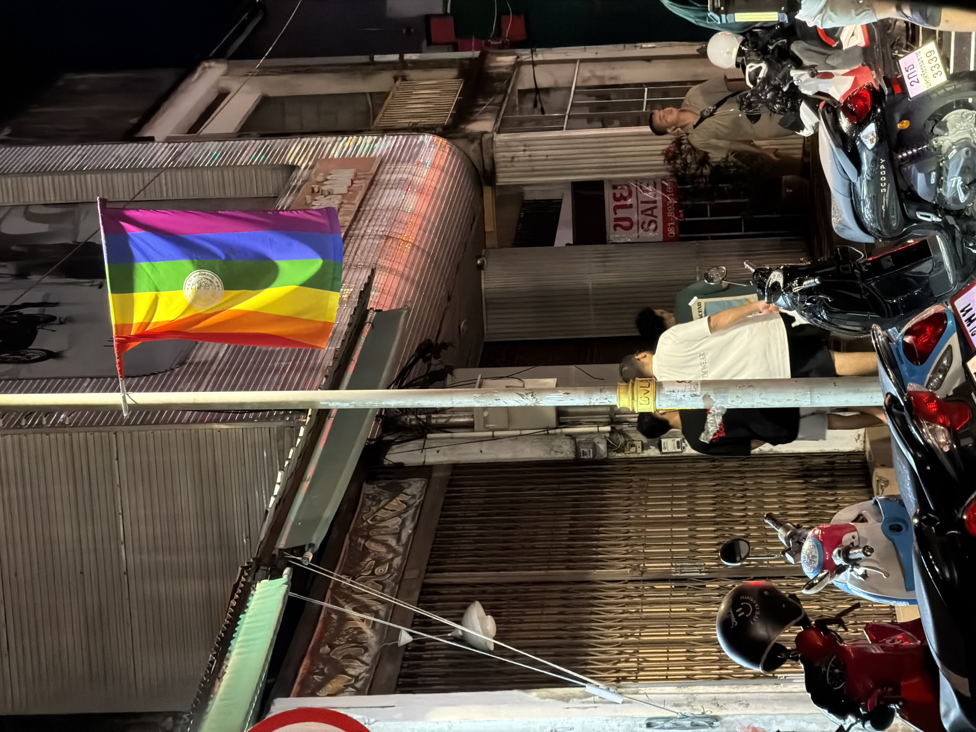In the city of Phuket City Municipality, Thailand, at night, a pride flag hangs from a pole on a street. The flag, featuring the rainbow colors with a circular emblem in the center, is positioned in front of a building with a corrugated metal facade. Several motorcycles are parked along the street, and a few individuals are visible. One person stands near a red motorcycle with a black helmet. Another group of three people are standing further down the street, one wearing a white t-shirt, another a green t-shirt, and the third a black t-shirt. A sign with Thai script and "SALE" is visible on the building. The environment appears to be a commercial or residential street, with the building housing storefronts or residences.