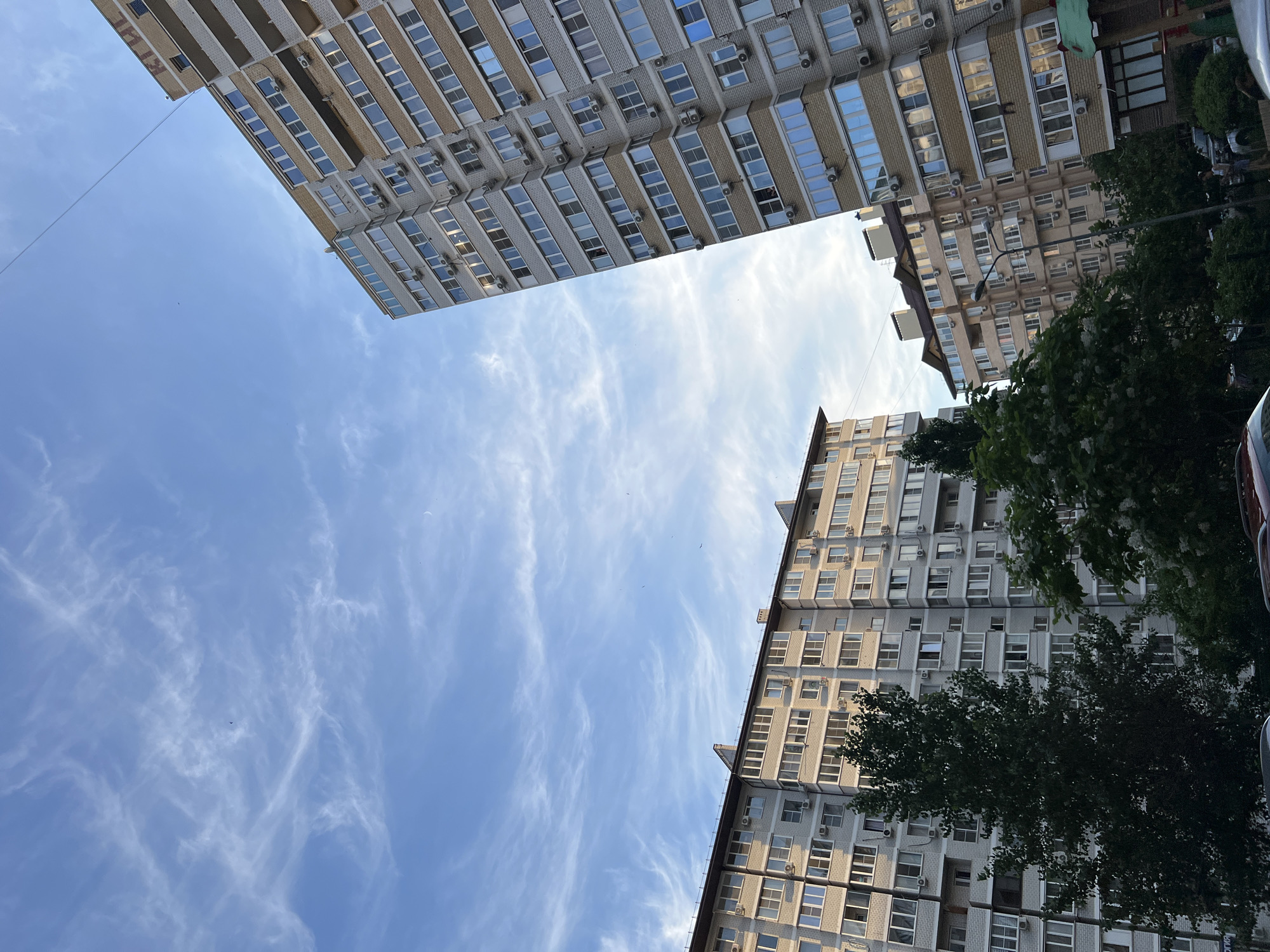 This media file captures an upward view of apartment buildings against a bright blue sky with wispy white clouds. The buildings are tall, with multiple stories and many windows. One building appears to be made of light-colored brick, while another has a more uniform, grey facade. Air conditioning units are visible on the exterior of the buildings. There are some trees with dark green foliage in the foreground and to the side, obscuring parts of the buildings. The perspective is from the ground looking up, giving a sense of scale and the height of the structures. The lighting suggests it is daytime, likely sunny given the bright sky. There is no visible text in the image. The scene depicts an urban environment, common in residential areas of cities.