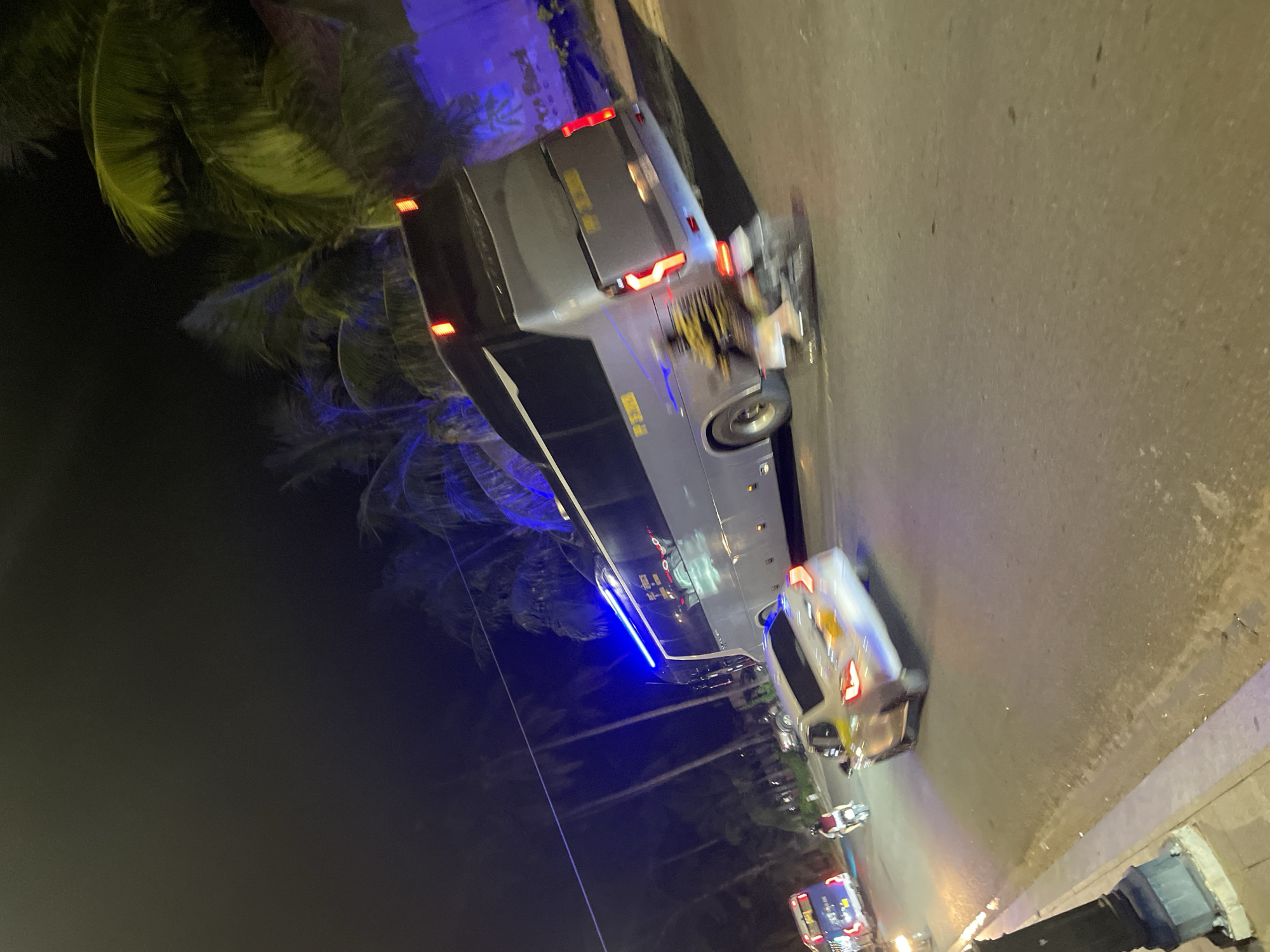 At night in Phu Thuy Ward, Vietnam, a silver car with its brake lights illuminated is captured in motion on a road lined with palm trees. A large gray tour bus is positioned to the right of the car, also in motion. In the background, a blue bus is visible, and a person is riding a motorcycle. The scene is illuminated by artificial lighting, including the headlights and taillights of the vehicles, and blue lights emanating from the tour bus and a structure in the background. The road surface appears to be asphalt.