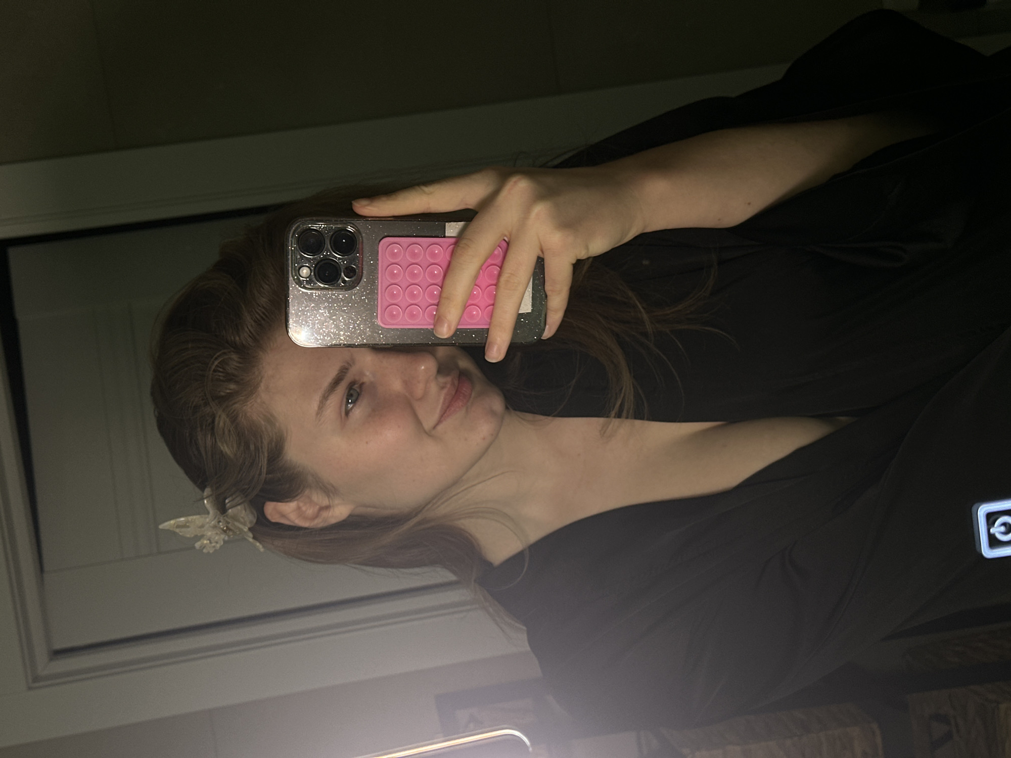 A person is holding a phone with a pink pop-it accessory in front of their face, taking a selfie. The phone has a glittery case. The person has fair skin, light brown hair, and is wearing a black garment. A butterfly-shaped hair clip is visible in their hair. The background includes a doorframe and wall, suggesting an indoor setting.