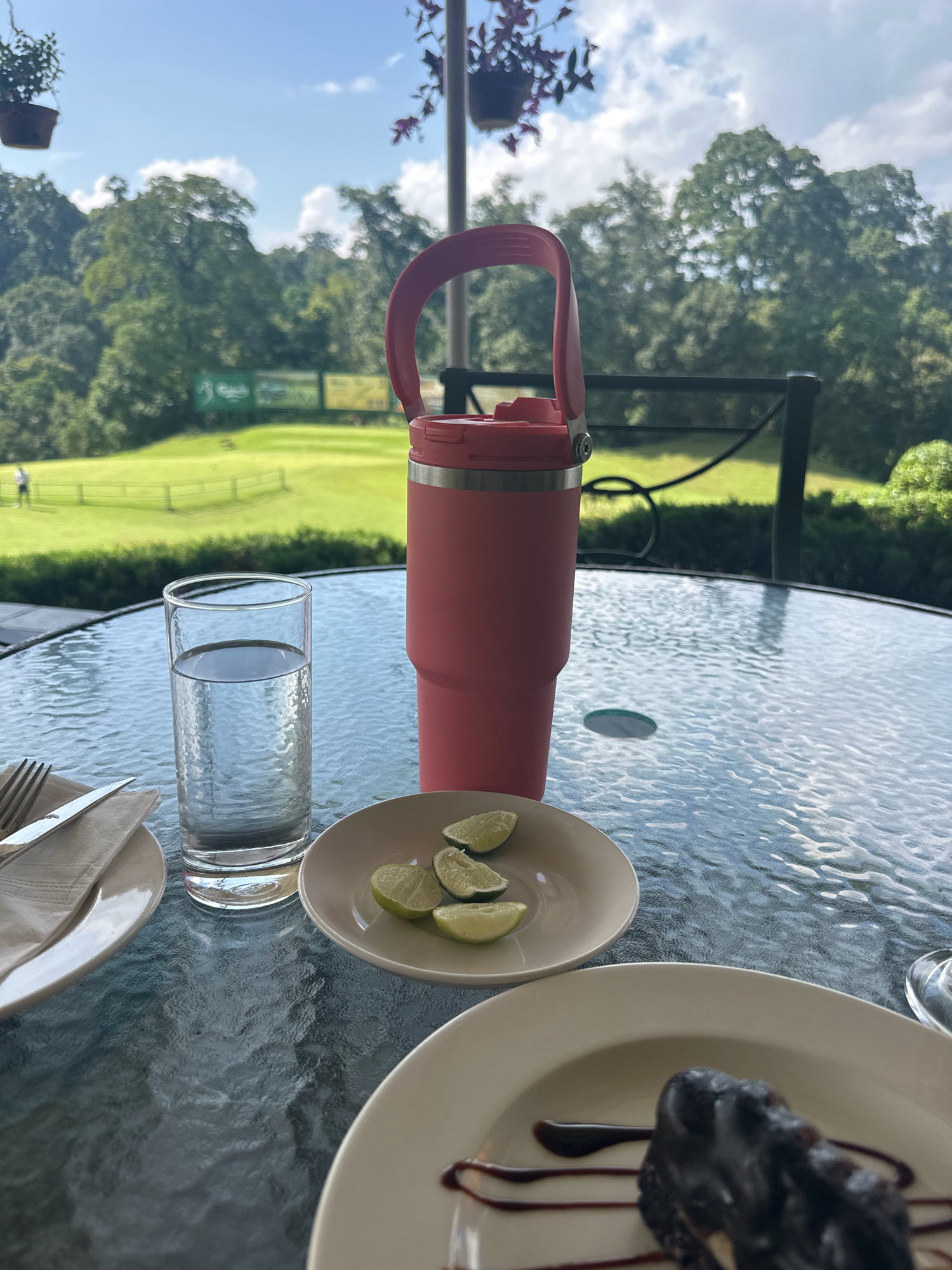 The image shows a table on a balcony overlooking a golf course. On the table are a glass of water, a pink tumbler, a small plate with lime wedges, and a dessert on a plate. In the background, there is a lush green golf course with trees and a blue sky with clouds. There are no people visible on the table or immediate surroundings, but one person is visible on the golf course in the distance. The scene appears to be a relaxed setting, possibly a restaurant or cafe with outdoor seating. The provided location is Kageshwori Manohara, Nepal.