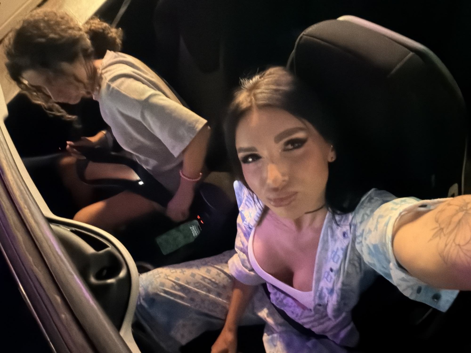 Two young women are in the backseat of a car at night. The woman in the foreground is looking at the camera with dark eye makeup and a light blue patterned shirt. She has a tattoo on her right arm. The woman in the background is wearing a light-colored shirt and looking down at her phone, which is on the car seat. A green phone is visible on the seat between the two women. The interior of the car is dark, with interior lighting casting a soft glow on the subjects. The car appears to be parked, as indicated by the stationary subjects. There are no visible landmarks to identify the specific location within the city of Dub, Montenegro.