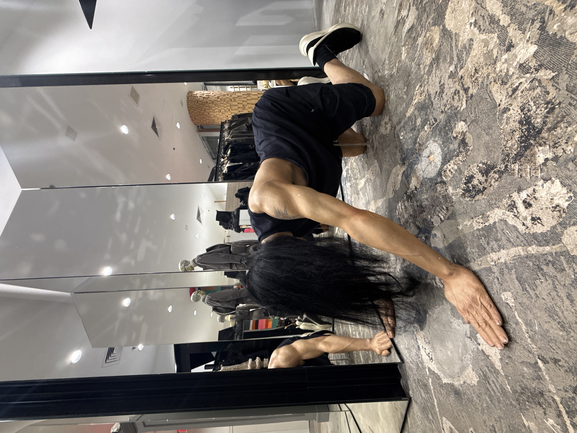 The scene depicts the interior of a high-end fashion retail store, specifically a Rick Owens boutique, likely located at 50 Wooster Street in New York City, United States of America.
A person with long, black, straight hair is positioned on a mottled grey, beige, and black patterned carpet, adopting a low, elongated posture on all fours. The individual wears a black sleeveless top, black shorts, black socks, and black and white athletic shoes. Visible muscular definition is present on the arms, with a faint tattoo or mark on the left bicep. The person's head is down, with hair cascading towards the floor, and hands are placed flat on the carpet in front.
The environment features multiple tall, black-framed mirrors reflecting overhead recessed lighting, white ceilings with air vents, and parts of the store interior. Reflections in the mirrors show at least two mannequins dressed in monochrome jackets, some with backpacks, standing in the background. Clothing racks displaying dark garments are visible further back. A distinctive, vertically oriented wooden pillar constructed from stacked wooden blocks stands in the mid-background, visible directly and in reflections. The walls are light-colored. The overall lighting is bright and even.