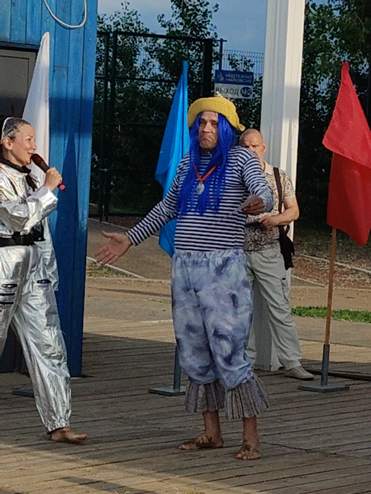 In an outdoor setting, three people are present on a wooden platform. The central figure is a man with blue wig and a yellow hat, wearing a striped shirt and patterned pants, with his arm outstretched. To his left, a woman in a silver, space-suit-like outfit is holding a microphone and speaking. Behind the man, another person stands partially visible, wearing a light-colored patterned shirt and light-colored pants, with a dark bag slung over their shoulder.
Two flags, one blue and one red, are visible behind the people, along with some trees and a fence. A sign with Cyrillic text is mounted on the fence. The lighting suggests it is daytime. The man in the blue wig and yellow hat has a stern expression, while the woman with the microphone appears engaged. The overall scene suggests an outdoor performance or event.