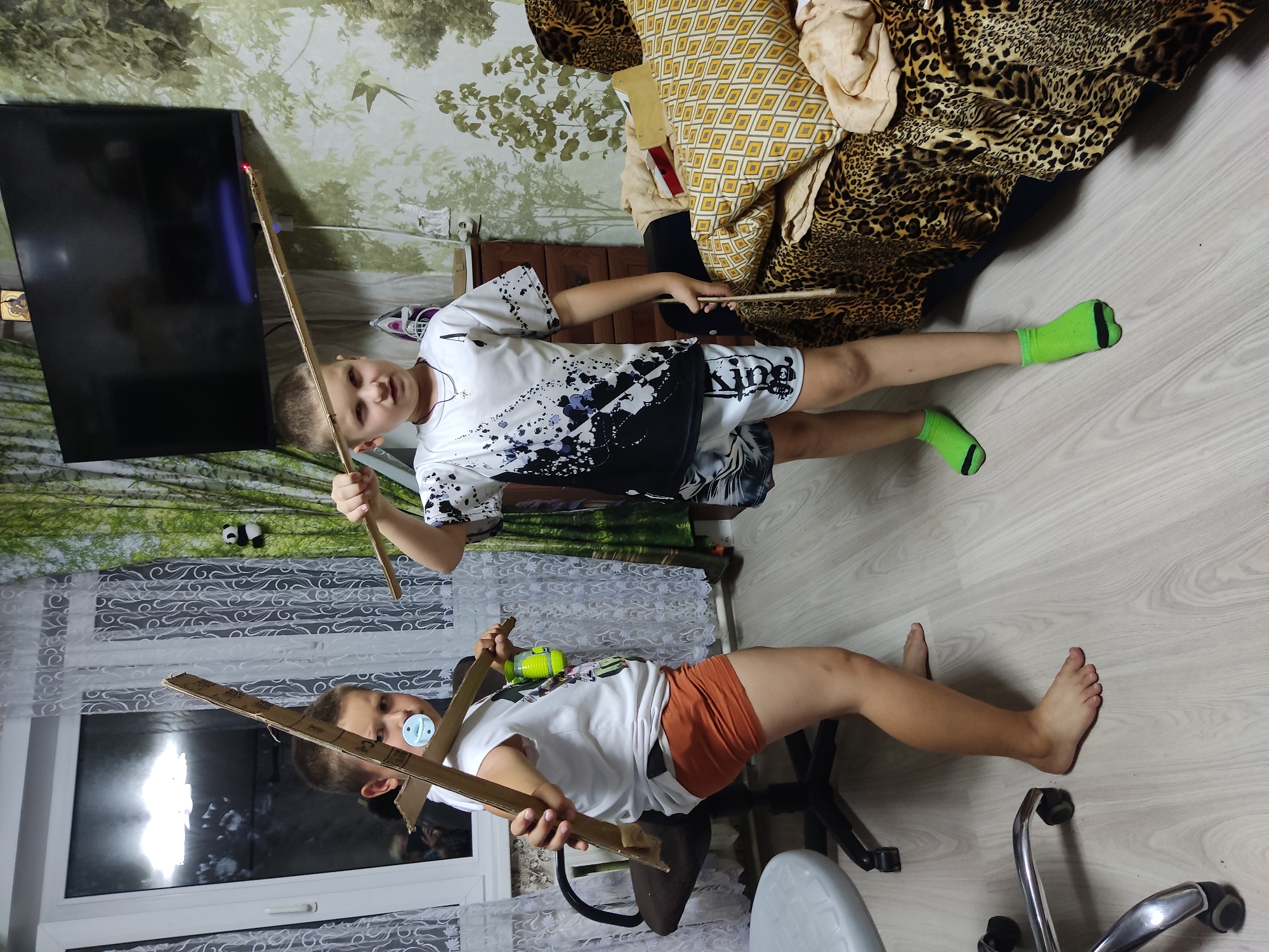 Two young boys are standing in a room, playing with homemade bows and arrows. The boy on the left is wearing a white t-shirt and orange shorts. He is holding a cardboard bow and arrow. The boy on the right is wearing a white t-shirt with a black and white abstract design and patterned shorts. He is holding a wooden stick with a red object at the end. The room has light-colored wood laminate flooring, a window with white lace curtains, and a wall with a nature-themed wallpaper featuring trees and birds. A large black television screen is mounted on the wall. A brown office chair and a cream-colored stool are visible. A sofa with leopard print fabric and patterned cushions is in the background. The scene appears to be in a residential setting.