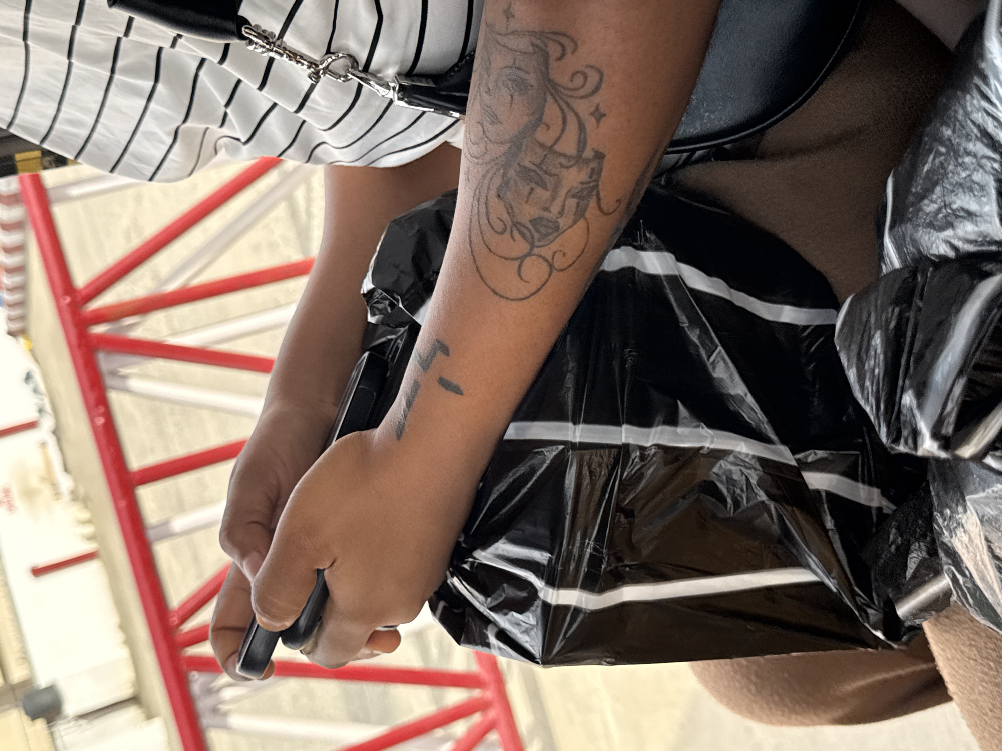 The image shows a person holding a black plastic bag with white stripes in their hands. The person has tattoos on their left arm. They are wearing a white shirt with black stripes and a black bag. In the background, there's a red metal structure. The location is Ado, Nigeria.