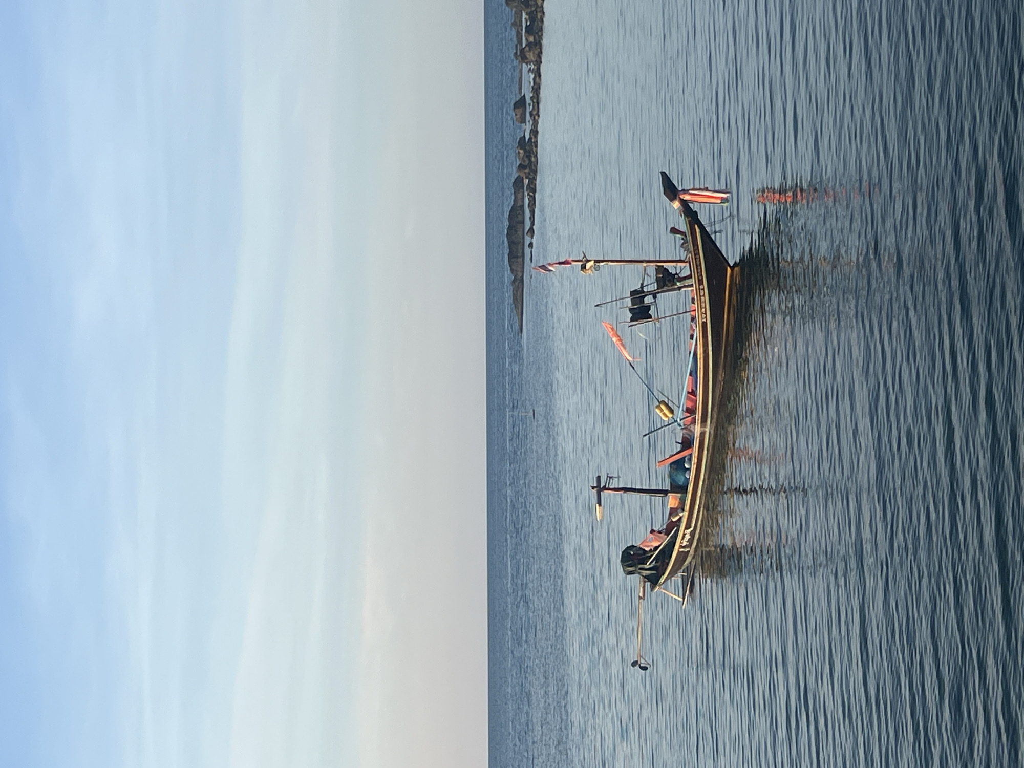 A long-tail boat is anchored in calm, blue water. The boat is brown with red and yellow accents and is equipped with a motor at the rear. It has a flag with red and white stripes flying from a pole. The boat is positioned in the lower half of the frame, with the ocean extending to the horizon. In the distance, a line of rocks is visible in the water. The sky above is a clear, pale blue, with a slight gradient towards a lighter shade at the horizon. The scene appears to be set during daylight hours, possibly early morning or late afternoon due to the soft lighting. There are no people visible in or around the boat. The location is Ko Pha-ngan District, Thailand.