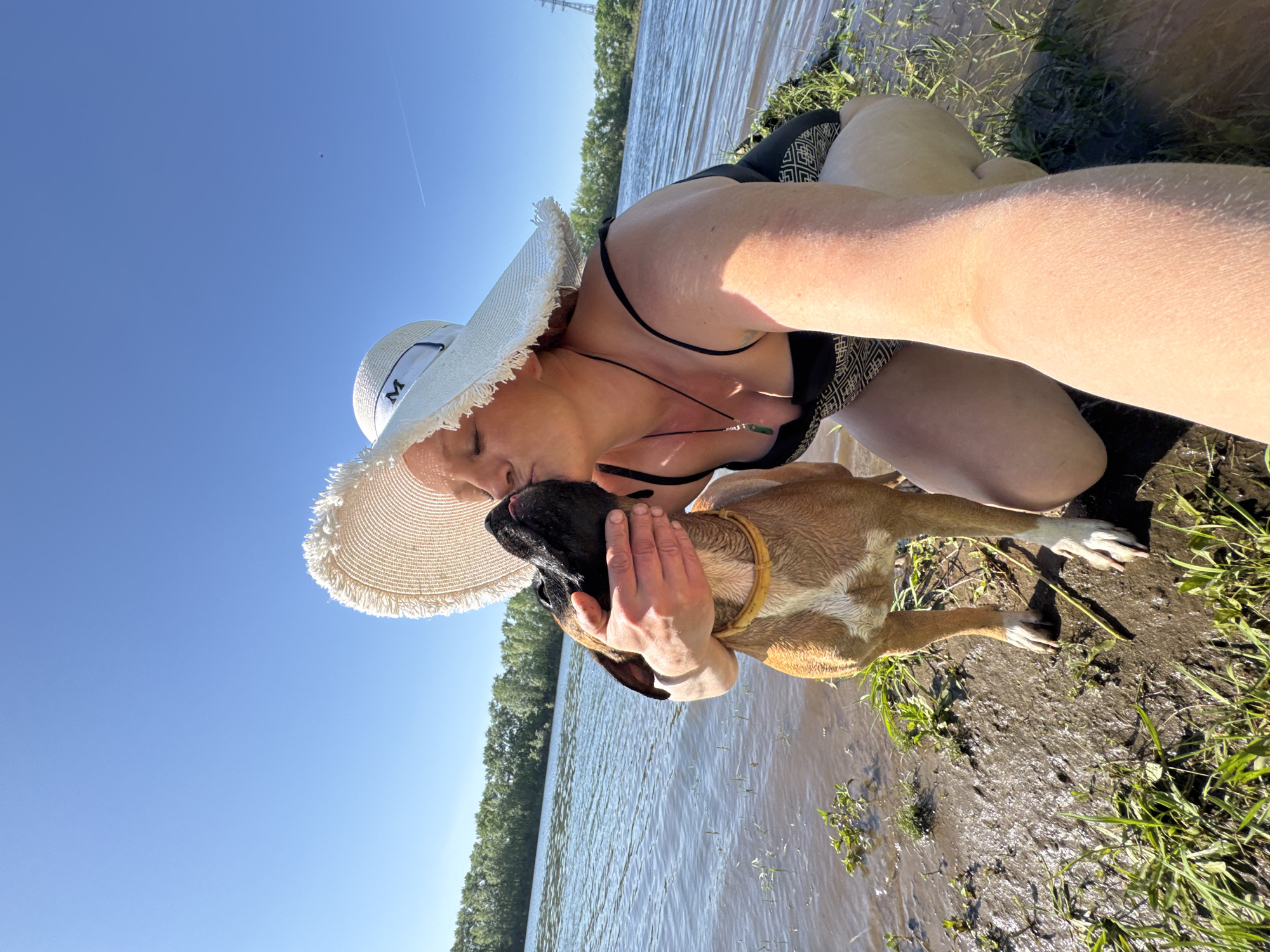 A light-skinned woman in a black patterned bathing suit, wearing a wide-brimmed white hat with the letter "M" on the front, is kneeling next to a body of water, possibly the Kama River in Chaikovsky, Russia. She is holding and kissing a fawn-colored Boxer dog with a black muzzle. The dog is wearing a yellow collar. The area around them is muddy with grass and vegetation. The sky is clear and blue. There is a line of trees on the opposite shore and a power line tower visible.