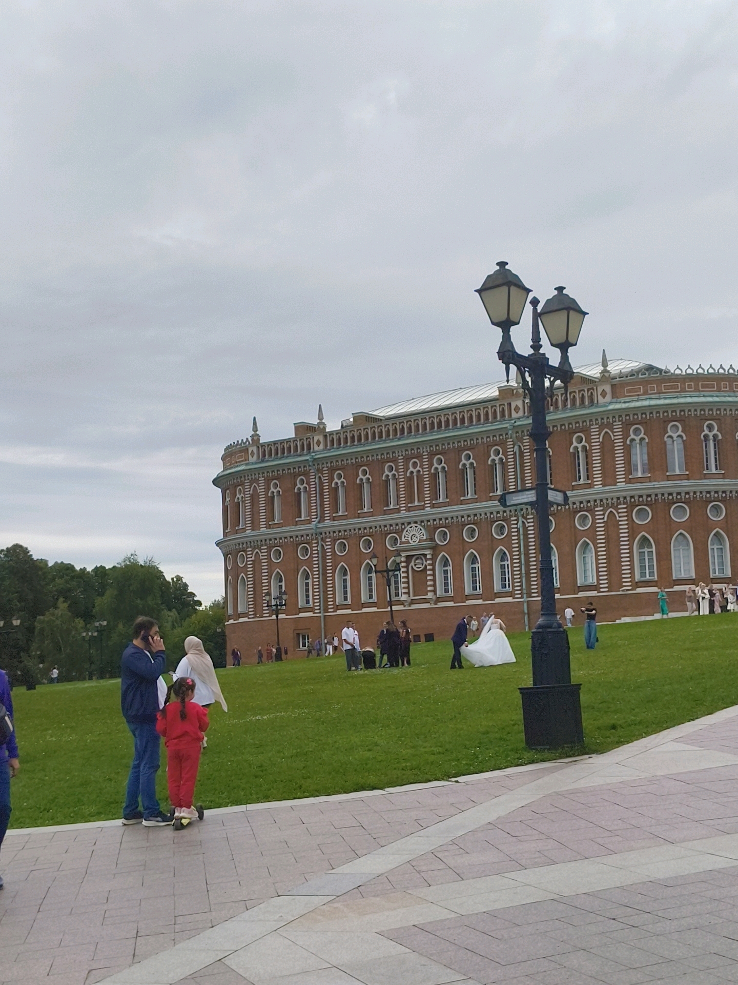 The image depicts a scene in Tsaritsyno Park, Moscow, Russia, with a large, ornate red brick building in the background. The building features multiple stories, arched windows with decorative white trim, and a green roof. In the foreground, a paved walkway is visible, leading towards a grassy area.
Several individuals are present on the grass. A man in a blue jacket and jeans is talking on his phone, standing next to a young girl in a red tracksuit who is riding a scooter. Further into the park, a wedding party is gathered. A bride in a white wedding dress and a groom in a dark suit are positioned centrally, with other guests in the vicinity, some of whom appear to be taking photos. A tall, black, double-headed lamppost stands prominently in the middle ground. The sky is overcast, suggesting a cloudy day. Lush green trees frame the left side of the scene.