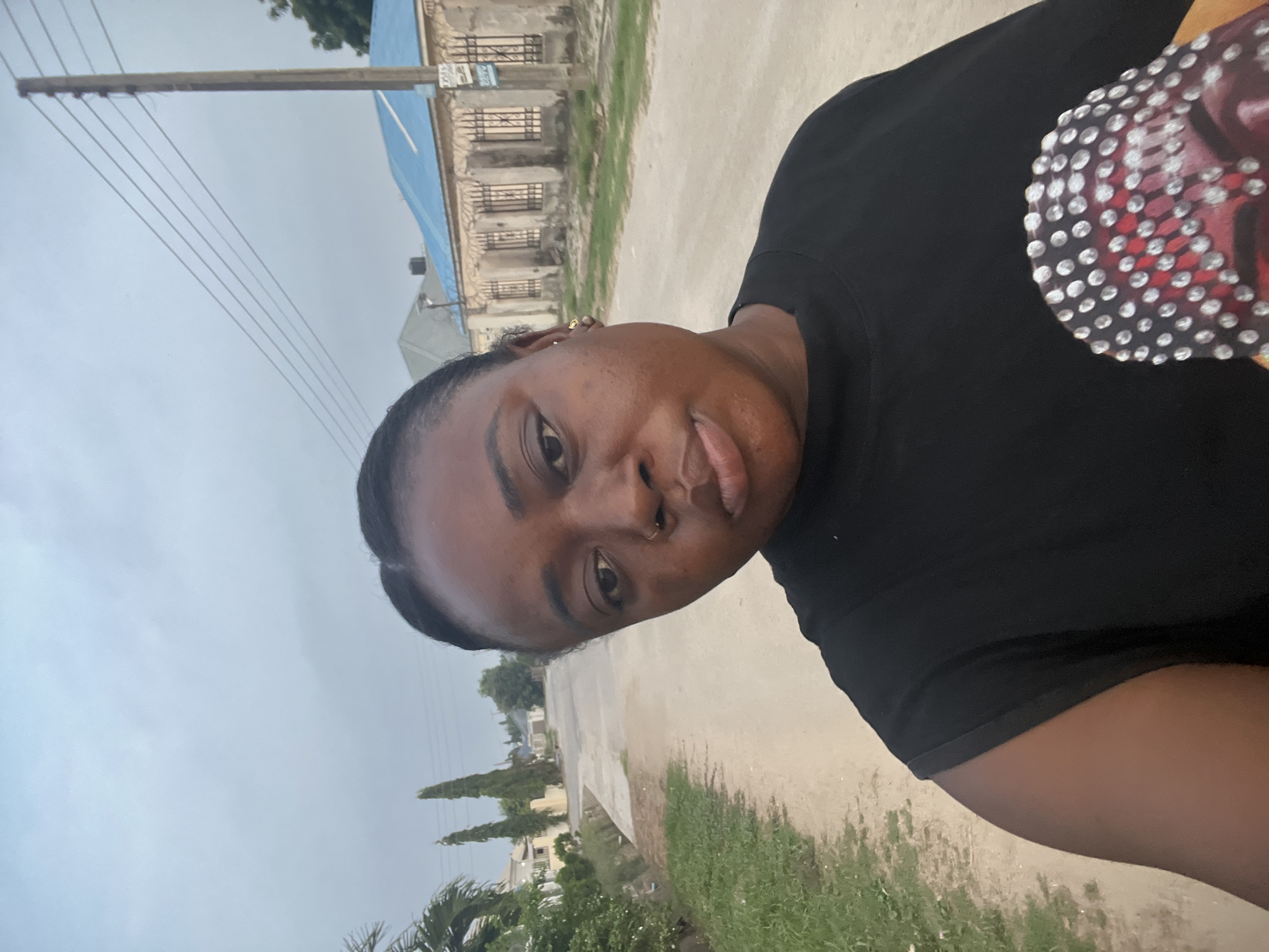 A woman is facing the camera on a road in Abuja, Nigeria. She is wearing a black t-shirt. A building with a blue roof and windows with bars is in the background. Power lines run along a pole next to the road. Green plants are visible along the edge of the road. The sky appears overcast.