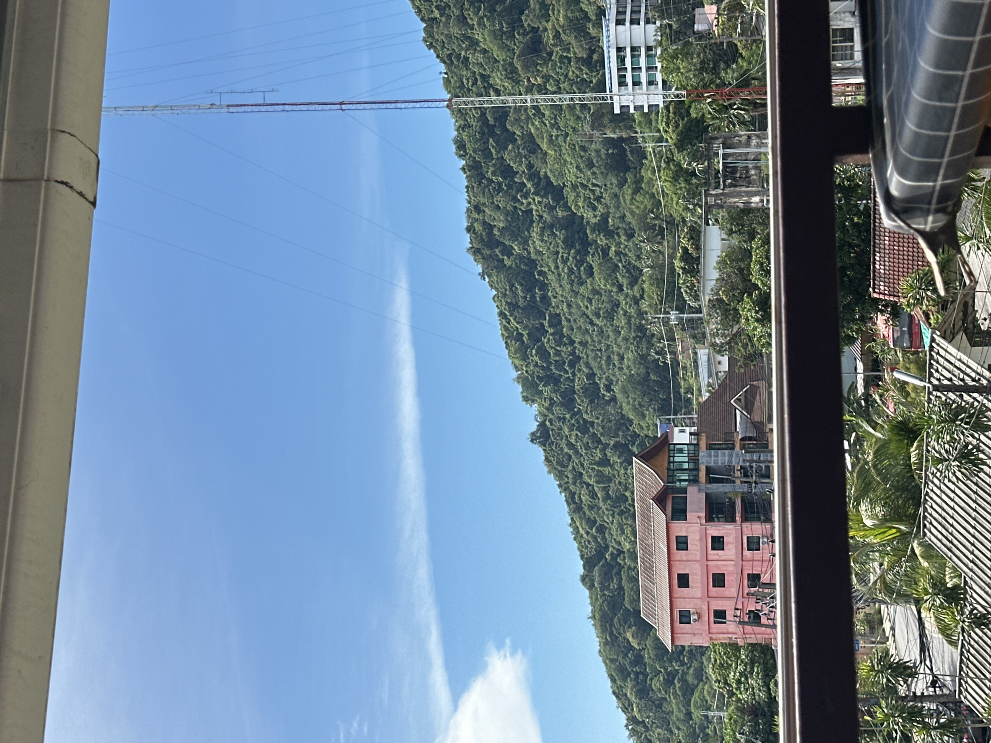 The image shows a view of a tropical hillside covered in lush green vegetation. A tall radio tower with cross-bracing structures extends upwards into the clear blue sky, with several power lines also visible. In the mid-ground, a cluster of buildings is nestled into the hillside. One prominent building is a multi-story pink structure with dark square windows and air conditioning units. Other buildings are visible behind it, including a white building with many windows higher up on the slope, and red-tiled roofs of residential homes. Palm trees and other tropical foliage are scattered throughout the scene, suggesting a warm climate. The foreground is partially obstructed by a dark metal railing and a white pillar, implying the photo was taken from a balcony or window. The bright sunlight and clear sky indicate it is daytime, likely mid-morning or early afternoon, with fair weather. The overall scene suggests a residential or tourist area in a hilly, coastal region. There is no discernible text within the image.