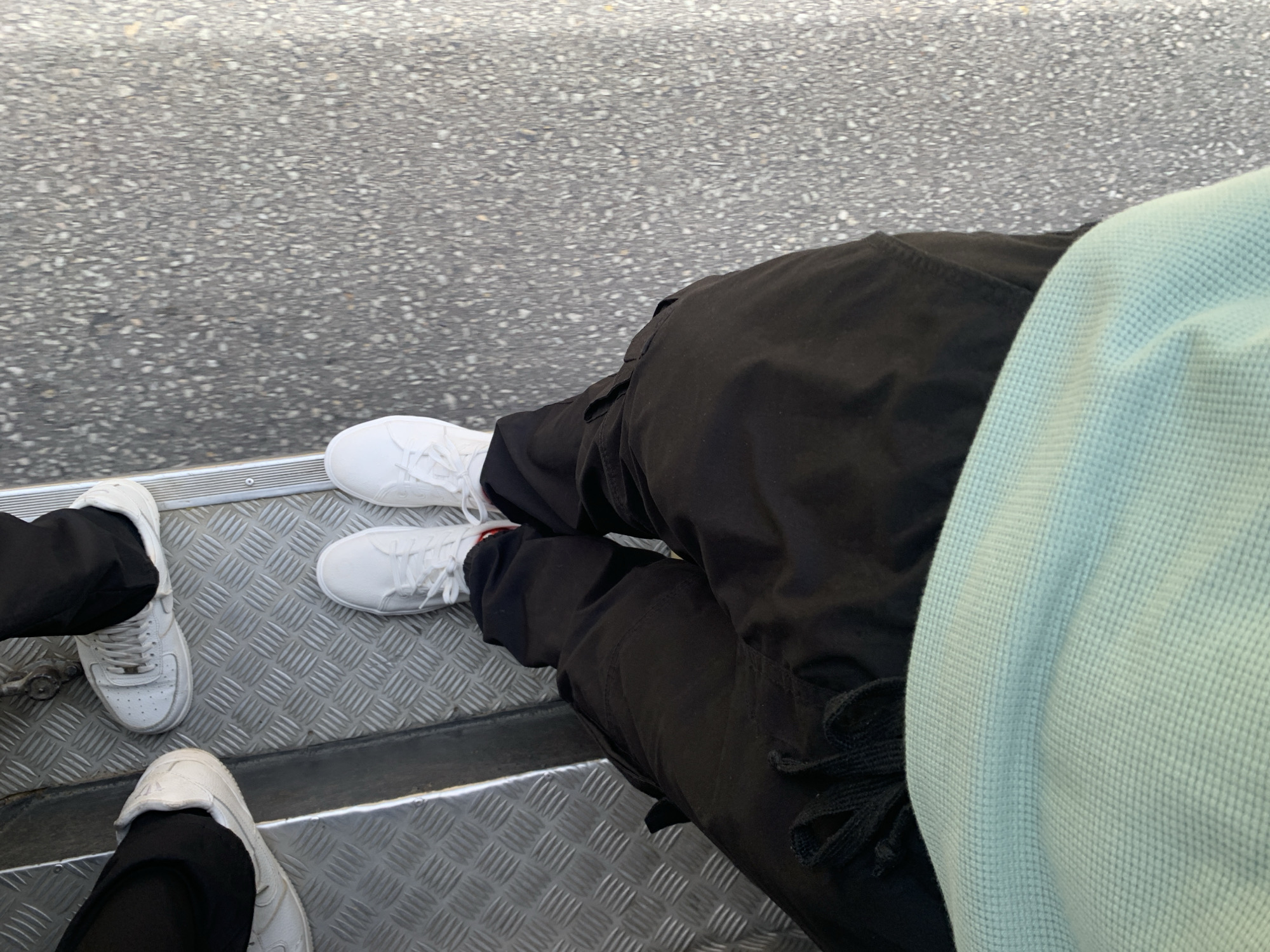 The image presents an overhead view of two individuals seated, likely within a vehicle or structure with metallic flooring. The person on the right wears black cargo pants and white sneakers, with a light blue waffle-knit garment partially visible. To the left, another person is visible with black pants and white sneakers. The ground visible outside the vehicle/structure is paved with asphalt. The exact location in La Asuncion, Venezuela cannot be determined.