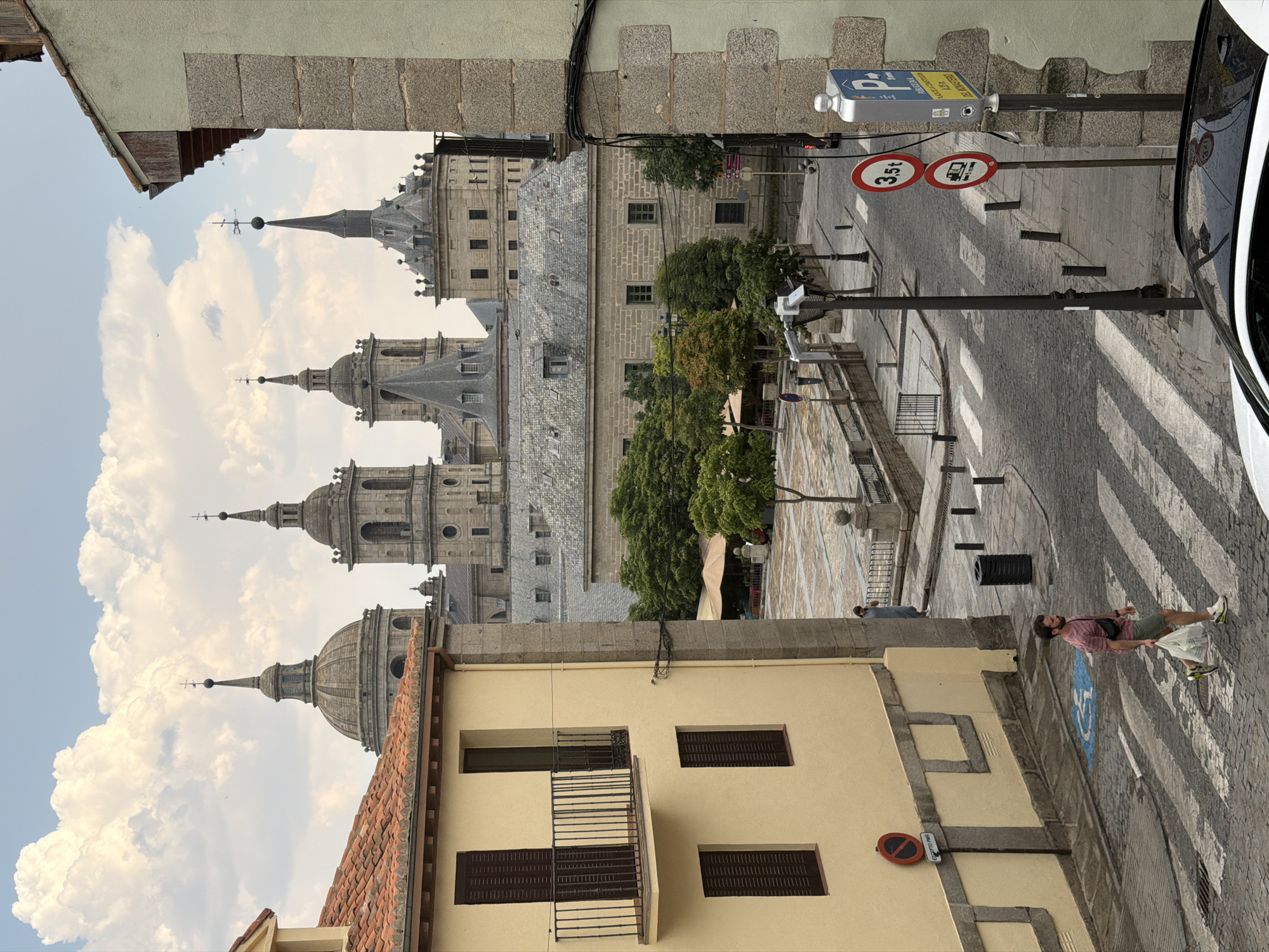 The image depicts a street scene in San Lorenzo de El Escorial, Spain, with the imposing Monastery of San Lorenzo de El Escorial visible in the background. In the foreground, a man walks across a pedestrian crossing, carrying a white plastic bag. He is wearing a striped t-shirt, khaki shorts, and athletic shoes. To his left, a yellow building with brown shutters and a balcony is visible. The street is paved with cobblestones, and there are several street signs, including a parking sign and traffic restriction signs. Trees and a market stall are present in the mid-ground, contributing to the urban landscape. The sky above is filled with dramatic clouds.
