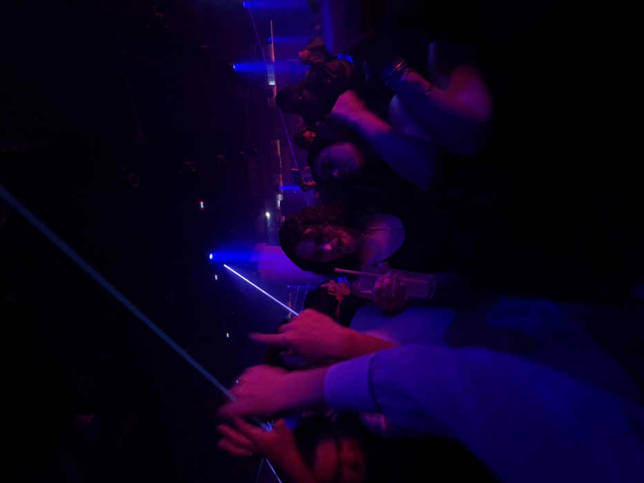 A dimly lit, vertically oriented shot shows people at a club or event. A laser beam diagonally cuts through the left of the frame. Hands point with fingers, and others are raised in the air. Someone holds a drink in a clear container. The lighting is primarily blue and purple, with dark shadows obscuring details. People appear blurred and in motion, conveying a sense of energy.