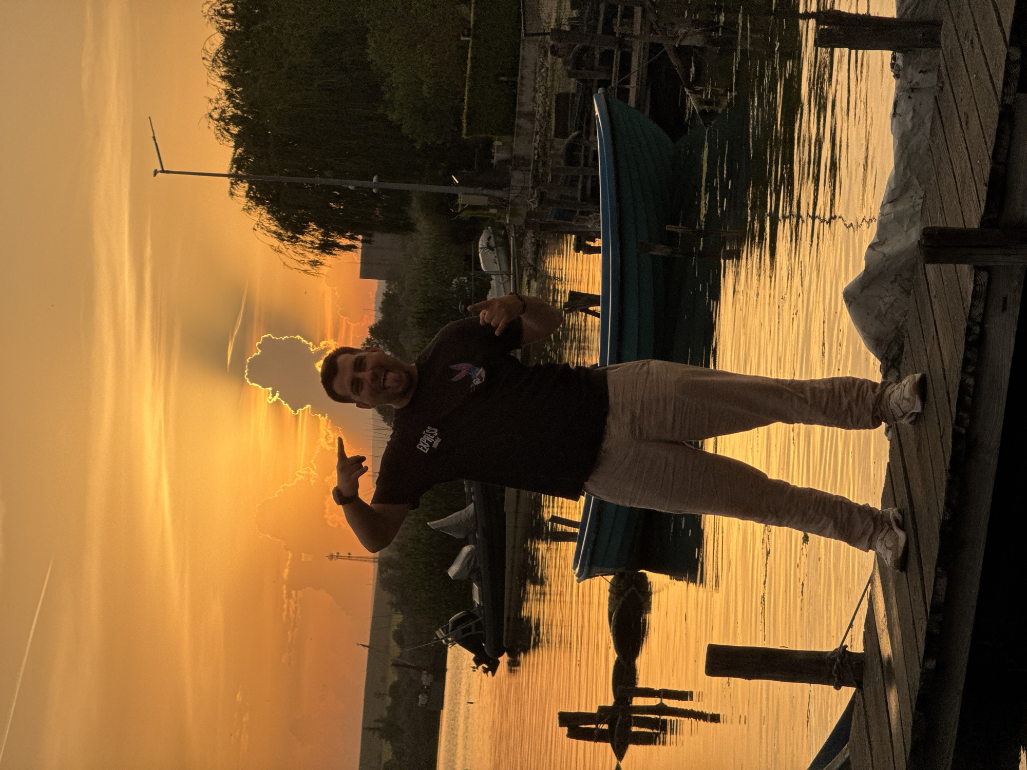 A man stands on a wooden dock beside a blue boat, with water reflecting a golden sunset behind him. The location is identified as Казашко, Bulgaria, evidenced by the landscape and architectural elements. He wears a dark t-shirt and light-colored pants and shoes. He makes hand gestures toward the sky. A structure with a sailboat mast stands in the background. Trees are visible along the distant horizon. The light creates a silhouette effect on some objects.