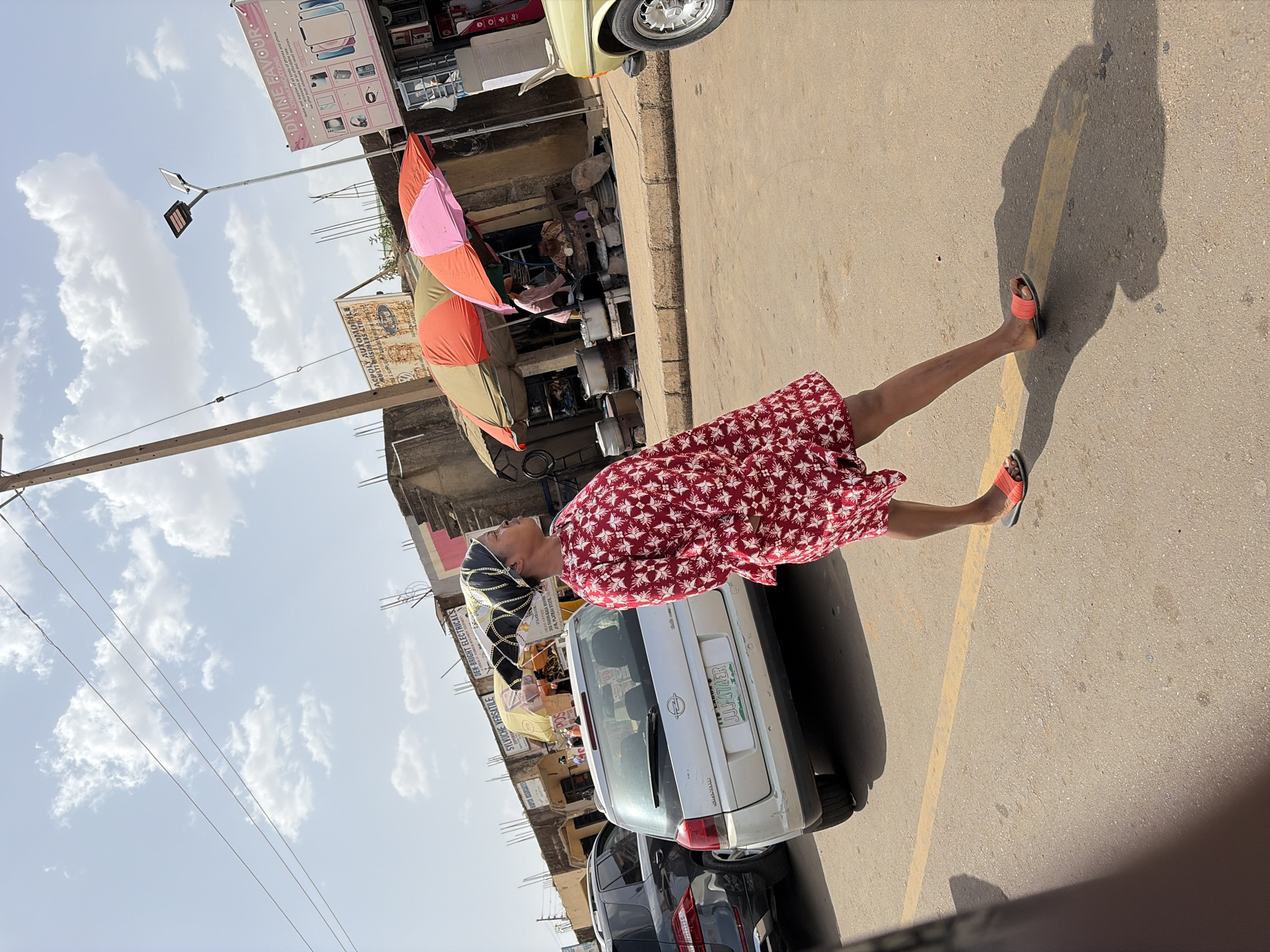 The image depicts an active urban street scene in Rafin Par, Nigeria, under bright daylight with scattered clouds and prominent shadows, suggesting midday. A woman is the central figure, seen walking or standing on a paved road, looking upwards. She wears a long-sleeved dress with a red and white geometric pattern, orange slip-on sandals, and a black head covering adorned with gold chains.
The background features several parked vehicles, including a white Opel/Vauxhall hatchback with the visible Nigerian license plate "JJJ-474ER JOS, PLATEAU STATE", and a partial view of a yellow classic car. Buildings line the street, housing various businesses. An individual holds a large, segmented umbrella (pink, orange, and brown) further down the road, with other people visible in the vicinity of the shops. Utility poles, overhead wires, and a modern street light structure are also part of the scene. The road itself has a yellow painted line.
Visible text on signs includes: "DIVINE FAVOUR • COMMUNICATIONS dealer and supplier of all kinds of phones and phone we offer both wholesale and retail services le rate", "SYLVUCHE VERSATILE", "DEN-BRIGHT ELECTRICALS", and a partially obscured sign reading "ACPOLY AUTOJAOTHLE MULTI BUSINESS LTL THE LADOS".