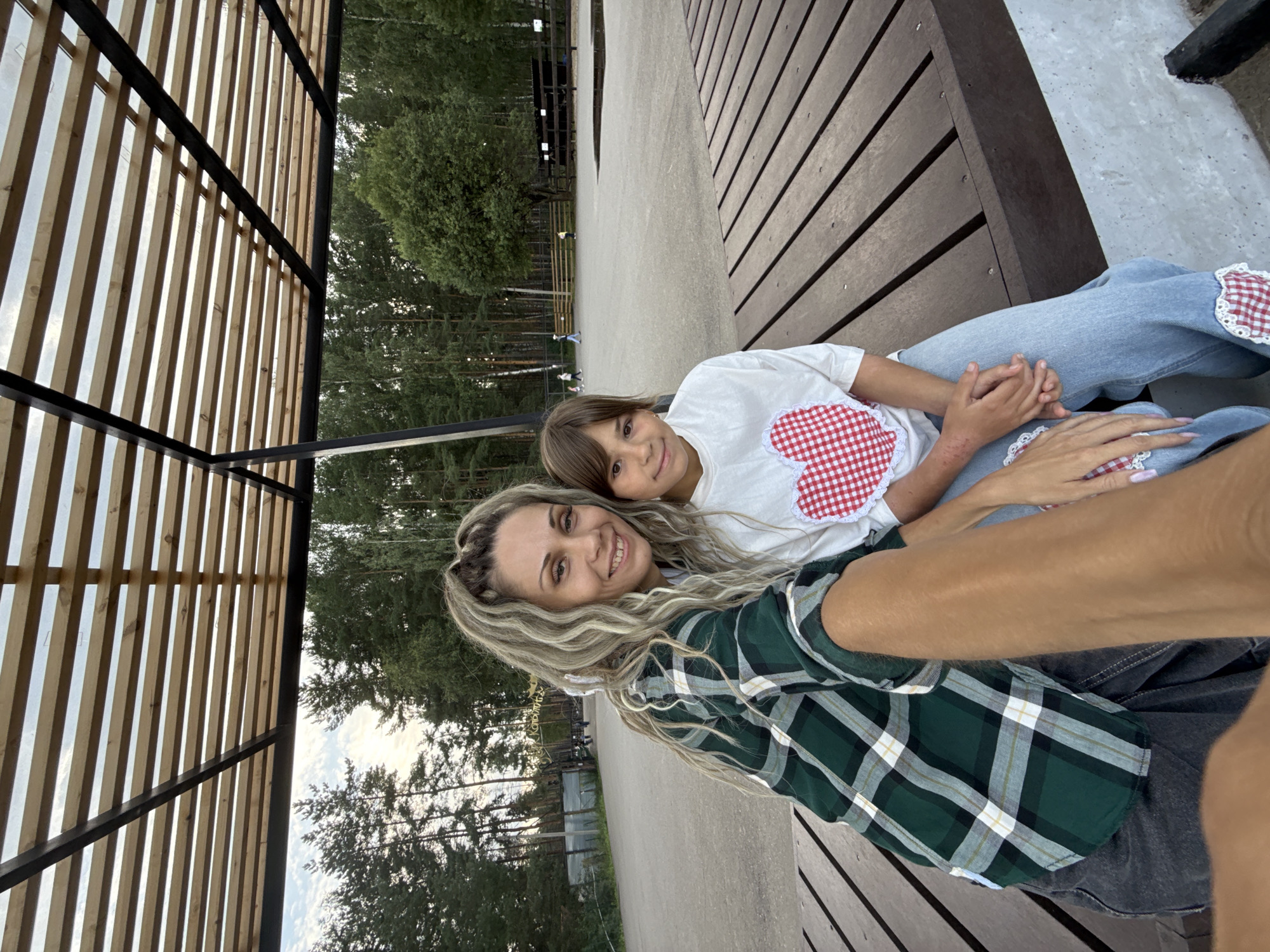 In Ignatovo, Russia, a woman and a young girl are seated on a wooden bench beneath a slatted wooden canopy. The woman, on the left, has long, blonde, wavy hair and is wearing a green and white plaid shirt over a white top and dark pants. She is holding a phone, taking a selfie, and smiling at the camera. The girl, on the right, has brown hair and is wearing a white t-shirt with a red and white checkered heart design and blue jeans. She is looking towards the camera with a slight smile. In the background, a paved area with scattered people, trees, and a wooden fence can be seen. The overall setting appears to be an outdoor recreational area or park.