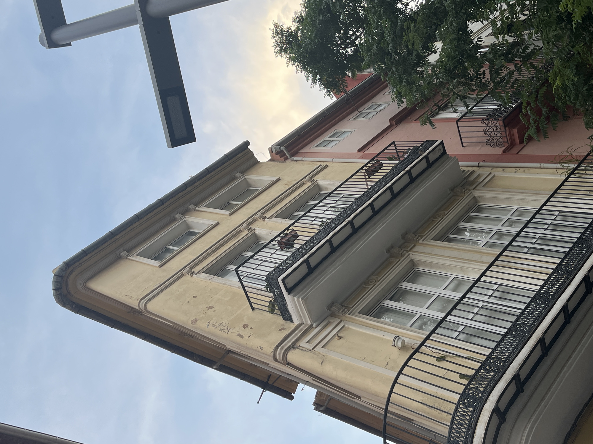 The image depicts a street-level view in what appears to be Valencia, Spain, based on the architectural style. The perspective is angled upwards, showcasing two buildings side-by-side. The building on the left is painted in a faded yellow and has multiple windows. It has a balcony with a black railing, on which a potted plant is visible. To the right is another building, painted in a pale pink or rose color, also with windows and a balcony. A tree with dense foliage obscures the top right corner of the image. In the top left corner, part of a modern street lamp is visible against a pale blue sky with hints of white clouds. The architectural details of the buildings suggest a historical urban setting.