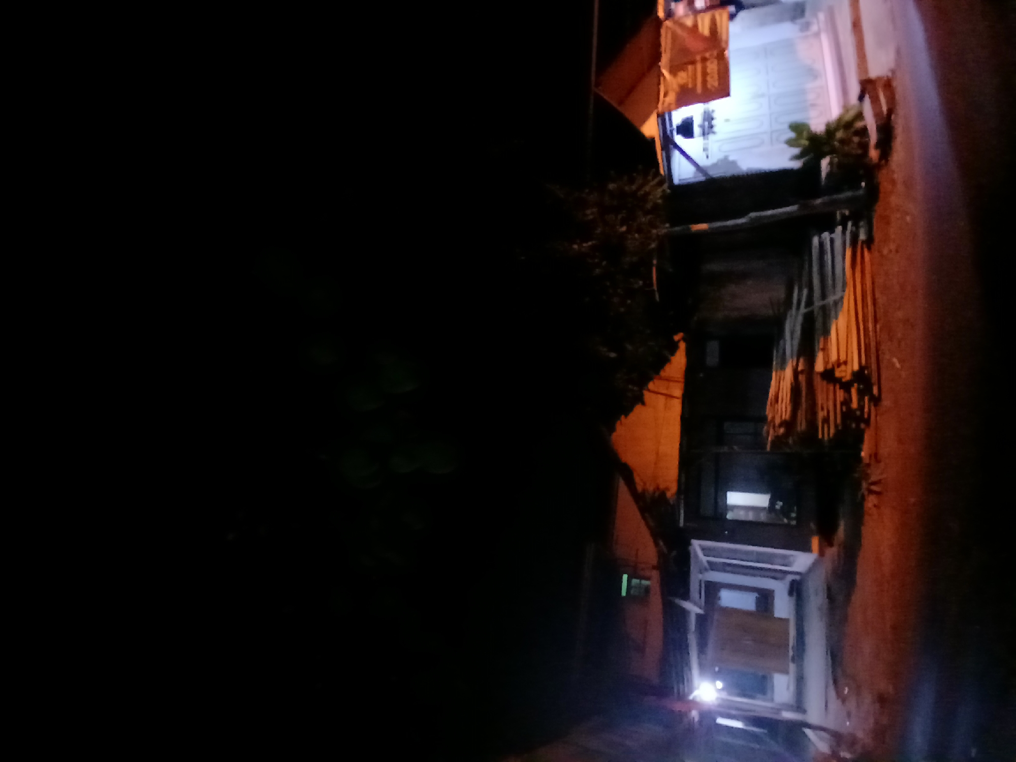 The image depicts a nighttime scene in Kademangan Kulon, Indonesia, featuring a row of buildings with exterior lighting. The structures appear to be residential or commercial, with one building showcasing a prominent orange-lit roof. In front of the buildings, a pile of lumber is stacked, suggesting construction or storage. The ground is a mix of dirt and pavement, with faint light illuminating the area. The sky is dark, with some indistinct green shapes visible at the top, possibly foliage or ambient light reflections. The overall impression is one of a quiet, illuminated street at night.