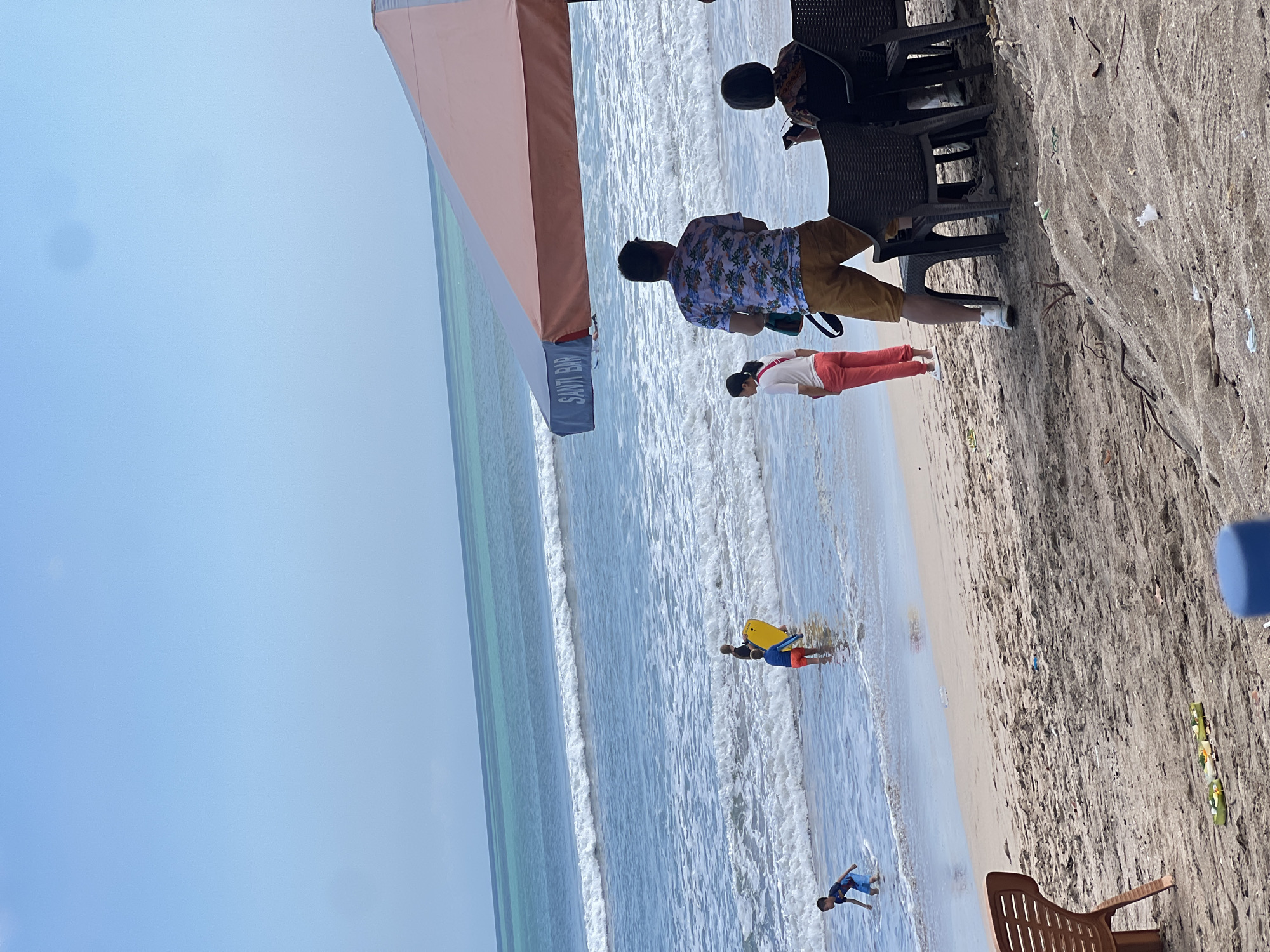 The image depicts a sunny day at Kuta Beach, Bali, Indonesia. In the foreground, several people are seated under a large umbrella. To the left, a brown plastic chair sits on the sand. Further down the beach, closer to the water, three children are playing in the shallow waves. One child is holding a yellow boogie board. The ocean has moderate waves with white foam. The sky is a clear, light blue. The sand is light brown and appears somewhat dry with scattered debris.