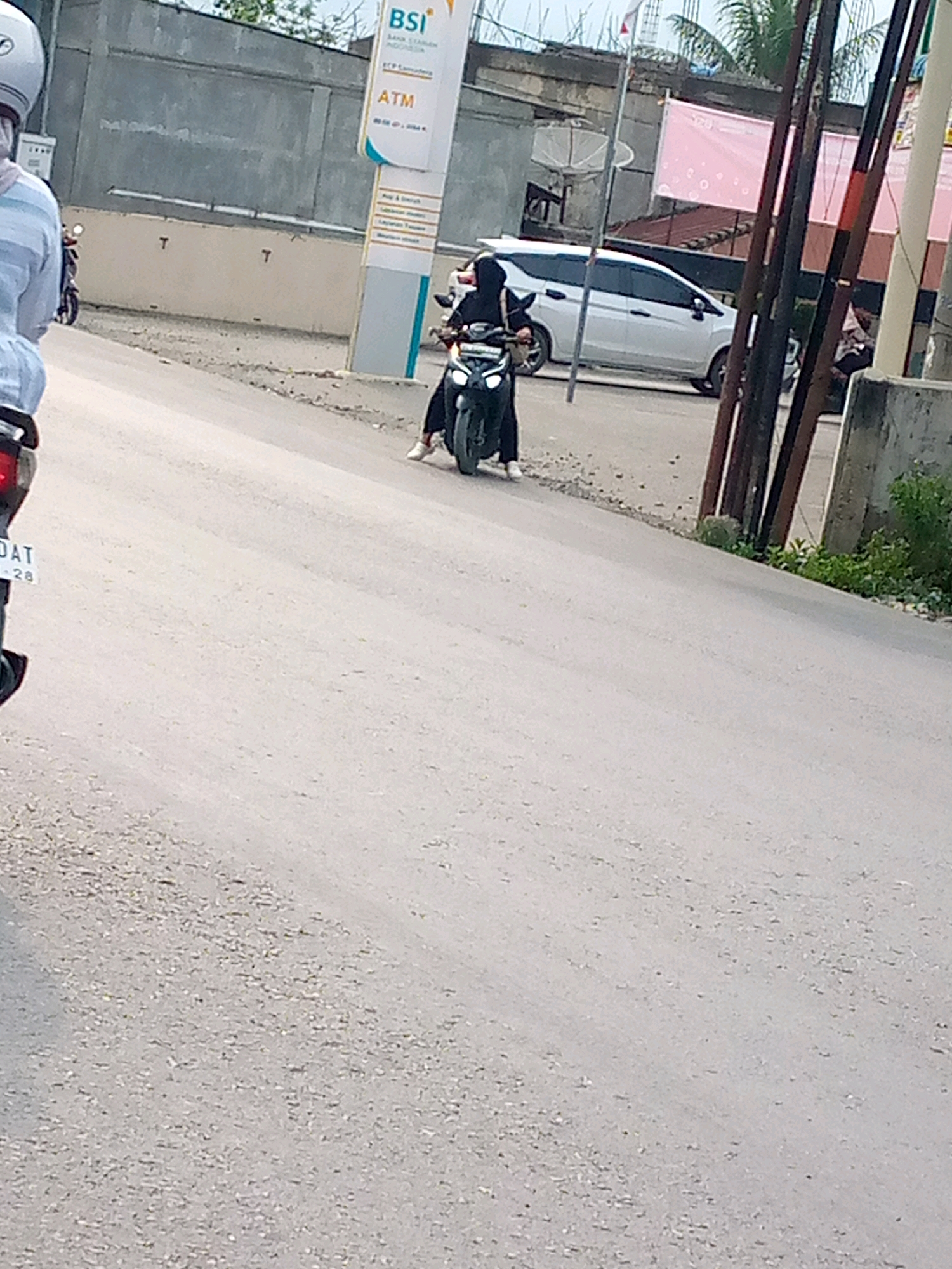 The image shows a street scene in Blangpeuria, Indonesia. In the foreground, the paved road is visible, with some gravel and debris scattered on its surface. On the right side of the road, there is a patch of green vegetation growing near a concrete structure and several utility poles. In the mid-ground, a person is riding a motorcycle, wearing dark clothing and a headscarf. The motorcycle is dark-colored. To the left of the motorcycle, a white van is parked or moving slowly. Further in the background, a building with signage is visible, including a sign for "BSI" and "ATM." There are also other structures and palm trees in the distance, suggesting an urban or semi-urban environment. A person on a scooter is also partially visible on the far left of the image.