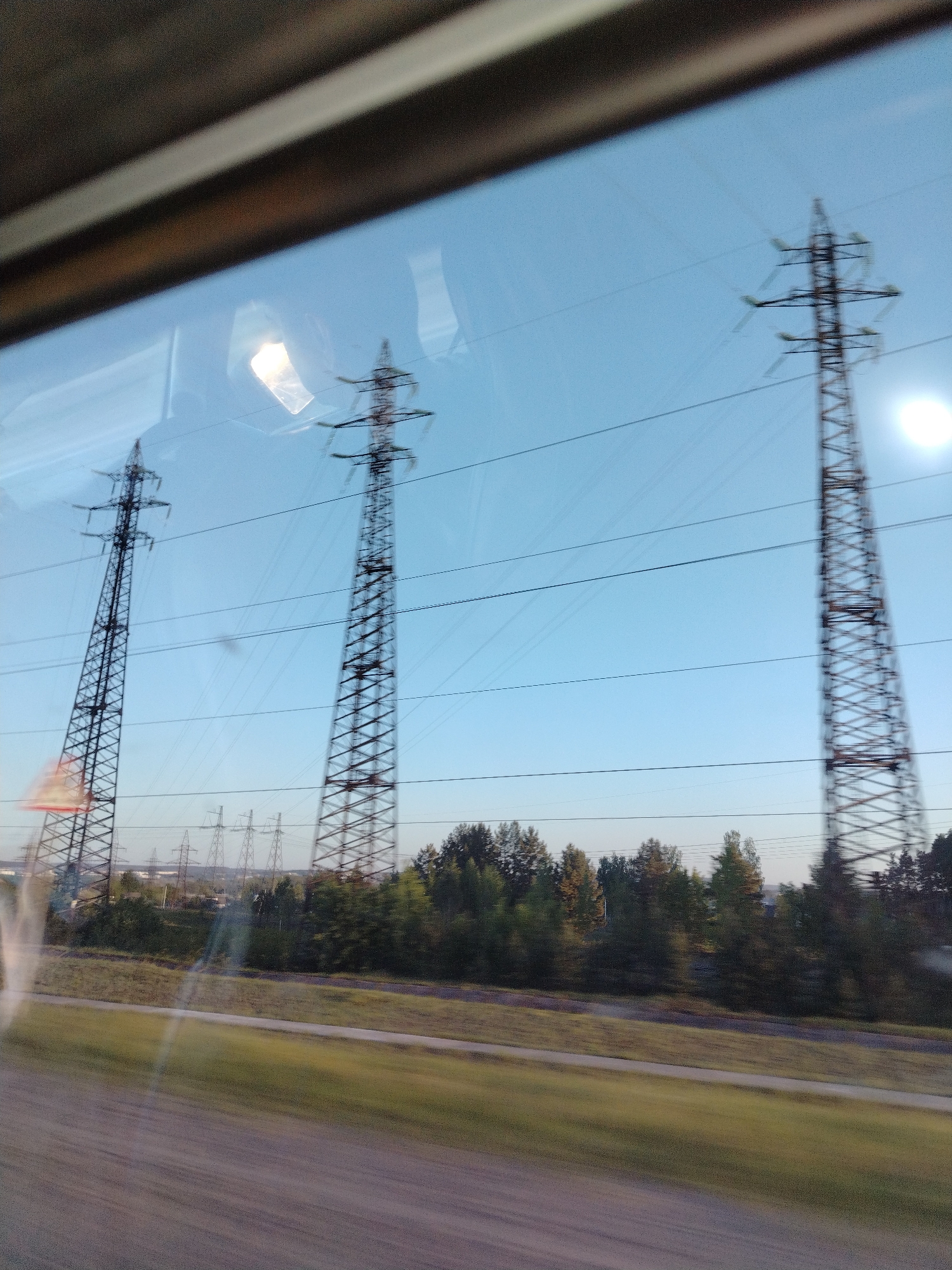 The image, likely taken from a moving vehicle through a window, shows a daytime view of high-voltage power lines and towers against a clear, light blue sky. The foreground includes blurred motion from the vehicle's movement, with a glimpse of vegetation and a grassy area outside. The power lines extend horizontally across the frame, connecting a series of metal towers in the distance. A line of trees is situated below the towers. The presence of power lines and towers suggests an industrial or suburban area. The metadata indicates that this image was taken in Noviy, Russia.