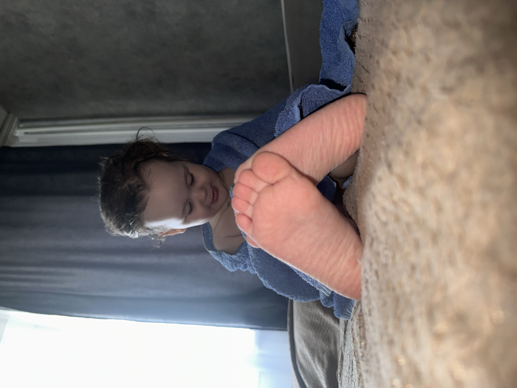 The image depicts a child lying on a bed, covered in a blue towel. The child is positioned near a window with grey curtains, suggesting an indoor setting. The location is Chaikovsky, Russia. The child's feet are prominently displayed in the foreground.