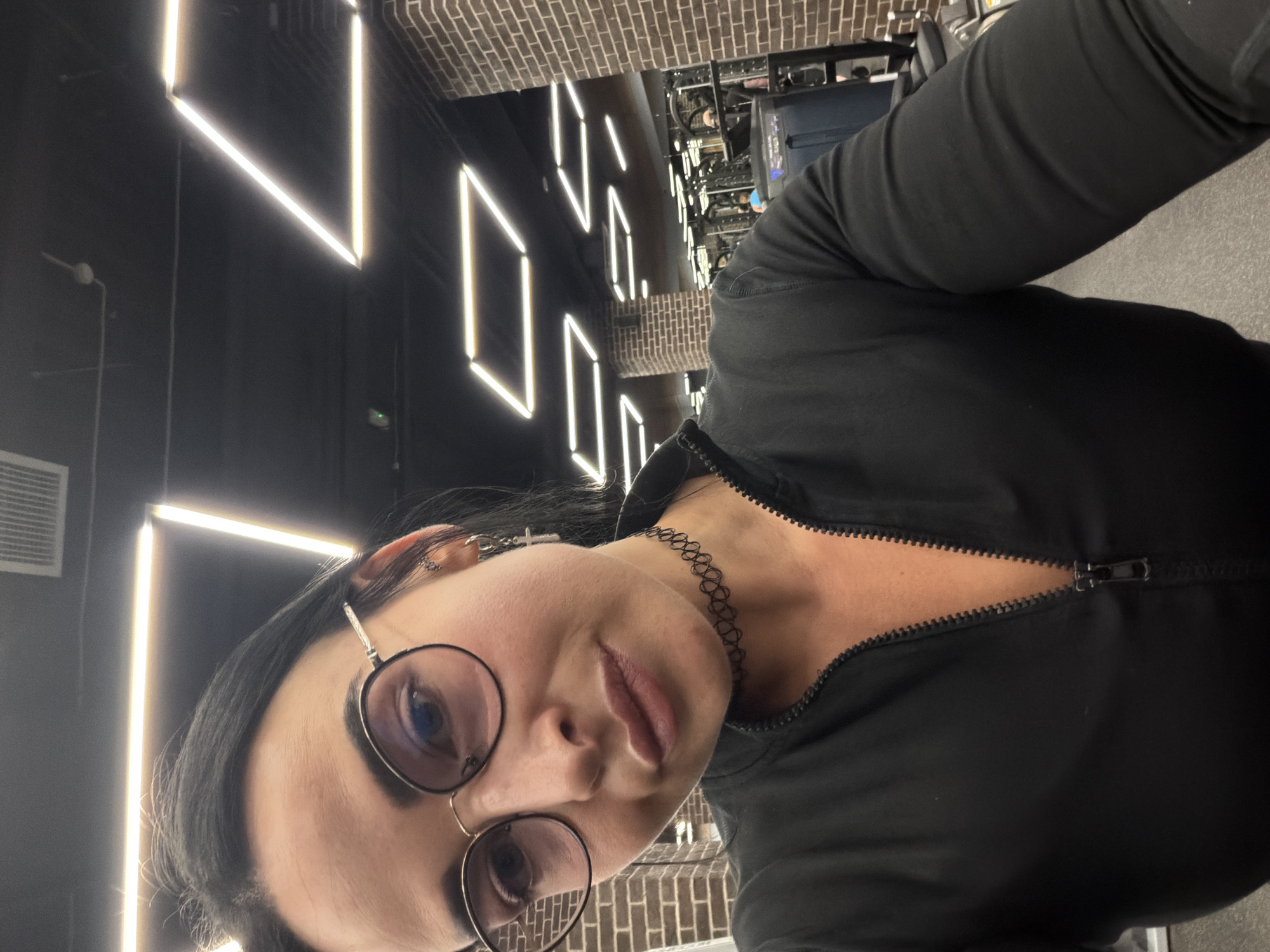 A female individual is depicted in a close-up selfie within a gym in Saint Petersburg, Russia. She has dark hair, wears circular-framed eyeglasses with metallic accents, a black choker necklace, and multiple earrings, including a silver cross-shaped dangle earring on her right ear. Her lips have a glossy appearance. She is wearing a black, long-sleeved athletic top with a partial front zipper, which is unzipped. The background reveals a modern gym interior with a black ceiling featuring illuminated linear and geometric LED light fixtures. Sections of the walls are constructed with exposed brick. Reflections in mirrors in the deeper background show gym equipment and other patrons. The visible flooring is grey.