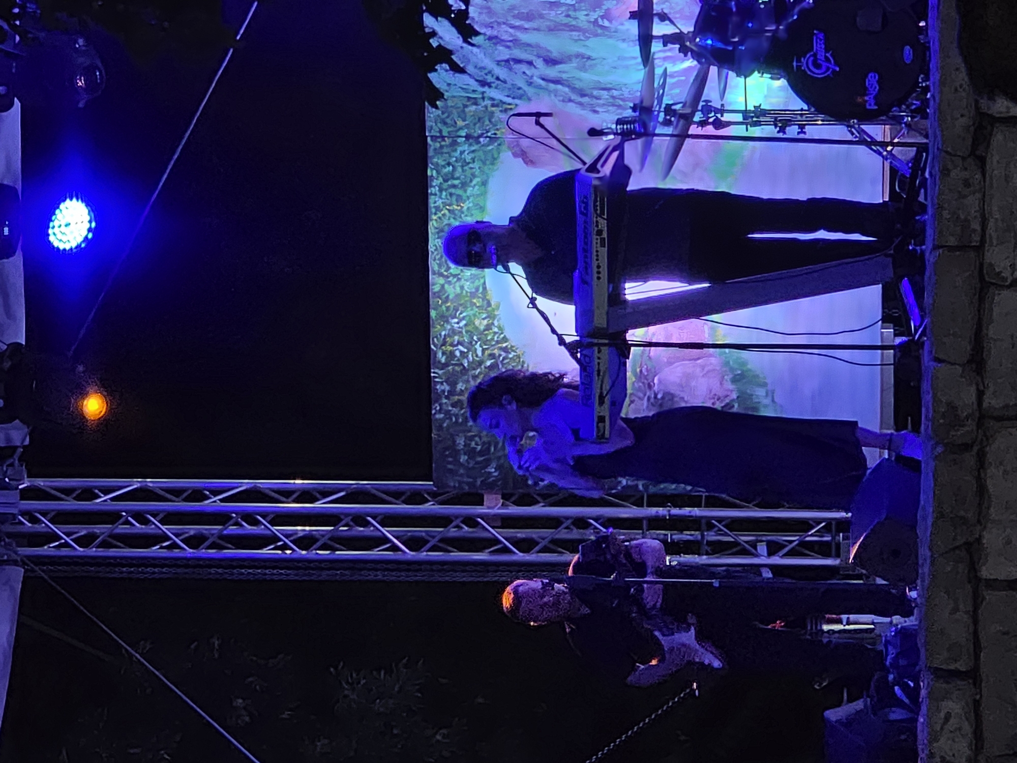 The image depicts an outdoor music performance on a stage at night. Three individuals are visible on stage. On the left, a man in a dark shirt is playing a white electric guitar. In the center, a woman in a dark dress is singing into a microphone while standing behind a keyboard. To the right, a man wearing sunglasses and a dark shirt is also behind a keyboard, with a drum set visible to his right. The stage is illuminated by blue and purple lights, and a backdrop with a scenic image of foliage is behind the performers. A metal truss structure is prominently featured on the left side of the stage. The performance appears to be taking place in an outdoor setting, possibly a park or an open-air venue, in Gara Oreshets, Bulgaria.