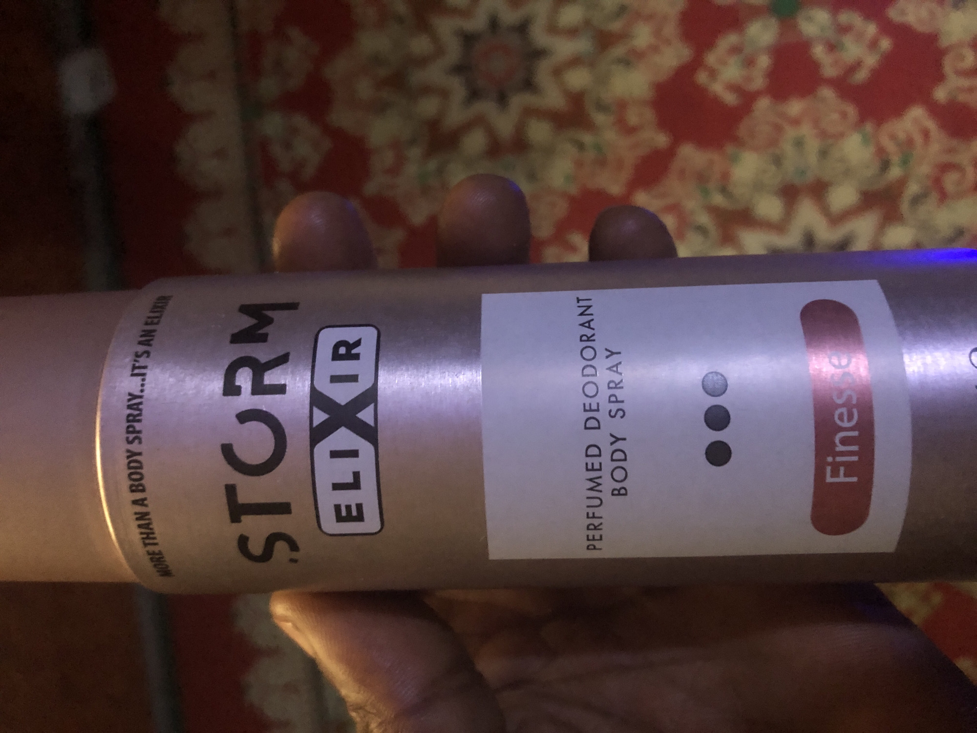 A person is holding a can of Storm Elixir perfumed deodorant body spray. The can is metallic and rose-gold colored. Text on the can reads "MORE THAN A BODY SPRAY... IT'S AN ELIXIR", "STORM", "ELIXIR", "PERFUMED DEODORANT BODY SPRAY", and "Finesse". The background is a red and yellow patterned fabric. The image is cropped such that the location in Kabong, Nigeria, is not visible.