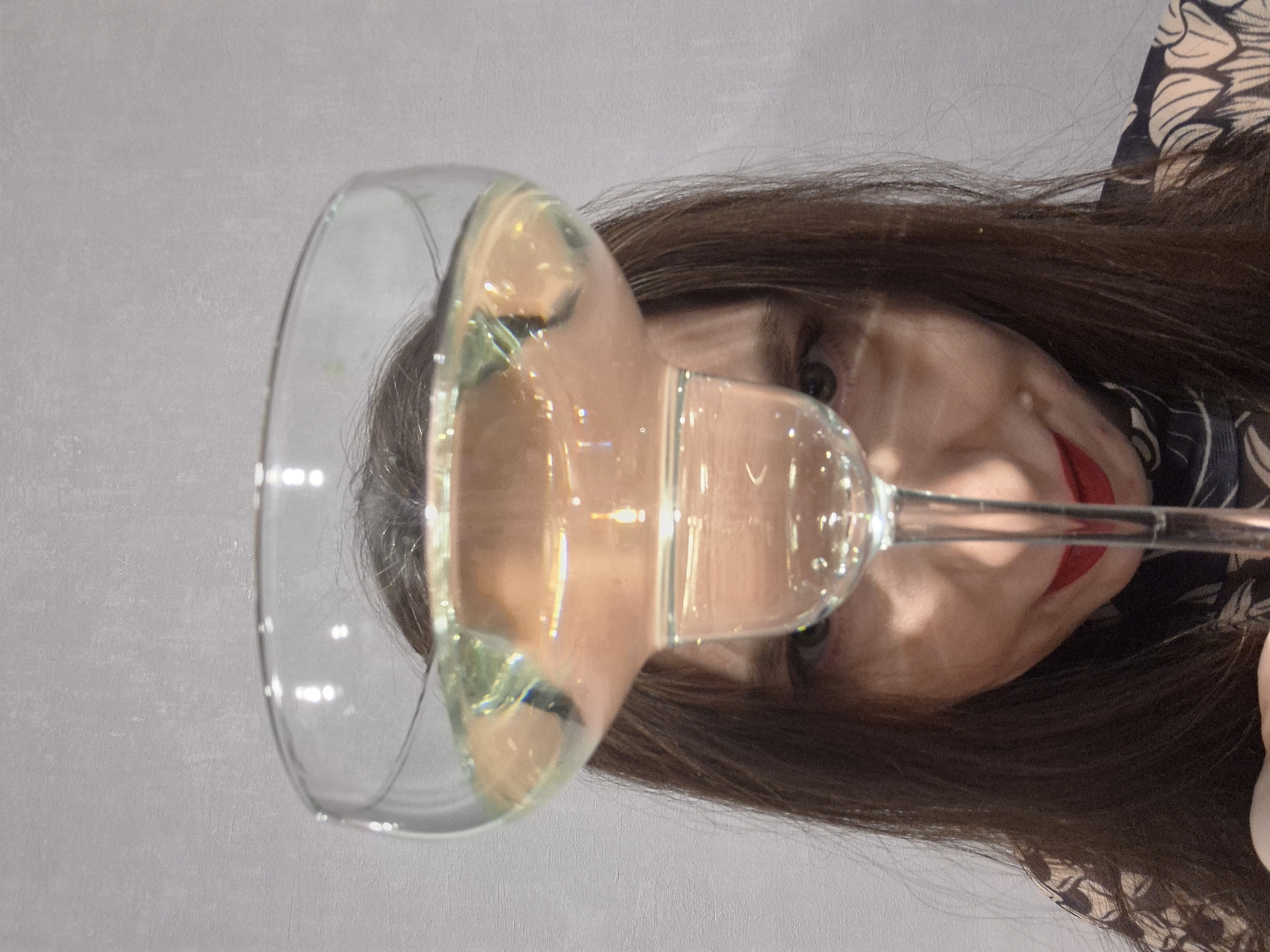 A fair-skinned woman with dark hair is positioned behind a clear glass containing liquid. She has red lipstick on. The background is a light gray wall. The woman appears to be holding the glass up in front of her face, partially obscuring it. The location and city of Chaikovsky, Russia are not definitively discernable from the provided image.