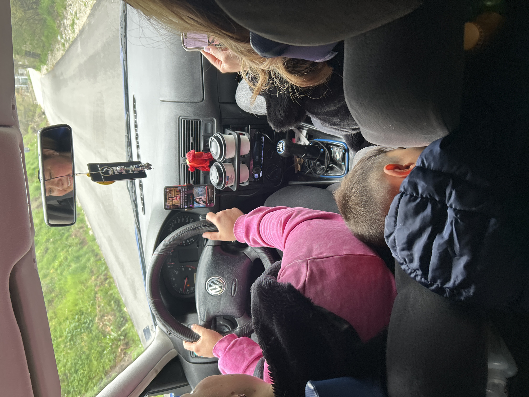 The image provides an interior view of a car, likely a Volkswagen due to the "VW" logo on the steering wheel, traveling on a narrow, paved road in Borinovo, Bulgaria. The perspective is from a rear passenger seat, looking towards the front.
In the driver's seat, a person wearing a pink long-sleeved top and a dark, fuzzy vest or jacket holds the steering wheel. To their right, in the front passenger seat, a person with blonde/brown hair is engrossed in their smartphone. A smiling adult face is reflected in the rearview mirror, suggesting a relaxed atmosphere.
The car's dashboard features a smartphone mounted in a holder, displaying what appears to be a video or social media feed. Below this, a cup holder contains two white cups. A red artificial flower is decoratively placed in an air vent, and the gear stick for a 6-speed manual transmission is visible. Further back, in a passenger seat, a person wearing a dark blue jacket is seen resting or sleeping.
Outside the vehicle, a winding road surrounded by green grass and trees indicates a rural or semi-rural setting. The bright natural light suggests it is daytime. The scene depicts a journey with multiple occupants, some engaged with devices, others resting.