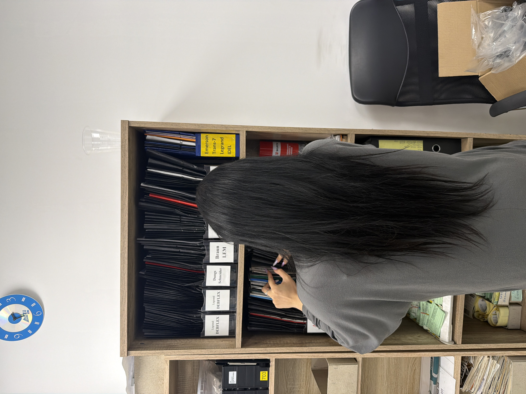 An overhead shot shows a person with long, dark hair standing in front of a wooden shelving unit. The shelves are filled with various items, including binders, folders, and boxes. The person is reaching into the shelves, appearing to search for something. A clock on the wall above the shelves shows the time. The overall environment appears to be an office or storage space. No specific landmarks or details indicating the city of Stara Zagora, Bulgaria are visible.