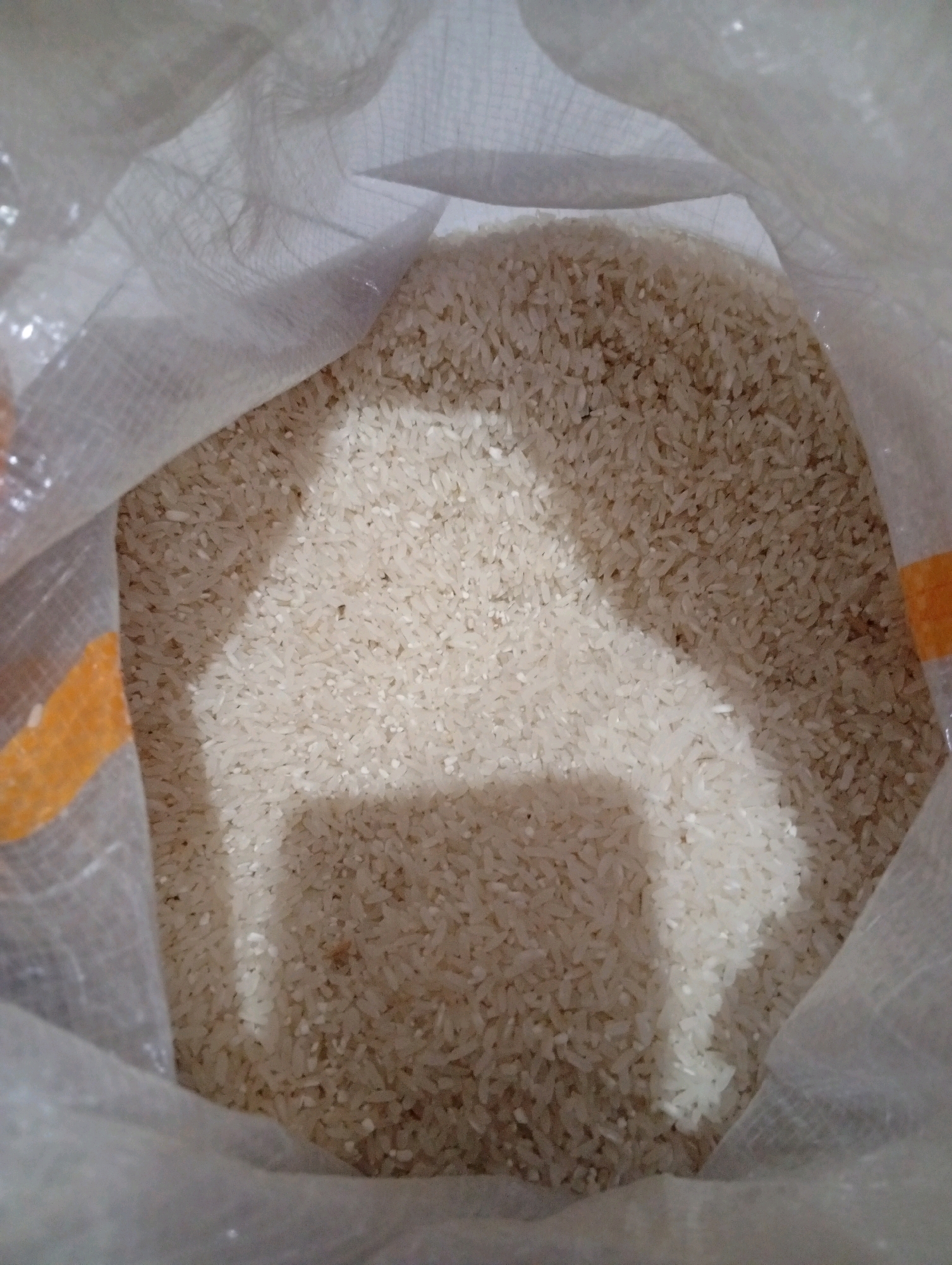 The image shows a close-up view of a white woven bag filled with uncooked rice. The bag is partially open, revealing the texture and color of the rice grains. Some areas of the rice are illuminated by light, creating shadows that add depth to the scene. The lighting suggests an indoor environment. No specific location in Tasikmalaya, Indonesia, can be determined from the image alone. There are no people or actions depicted.