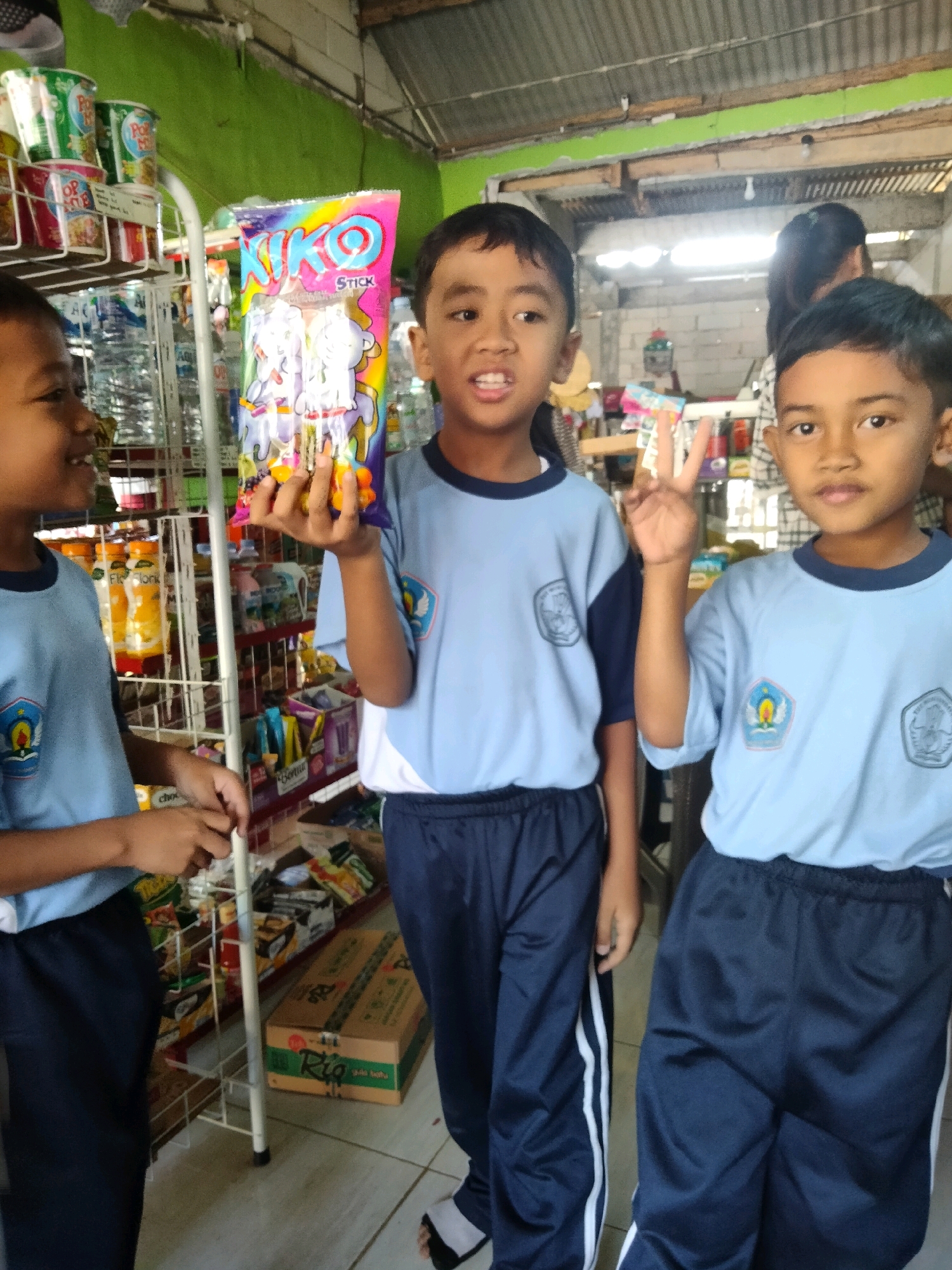 Three young boys, likely students based on their matching light blue shirts with school insignia and dark blue pants, are at a store in Sidorejo, Indonesia. The boy on the left, facing the camera in profile, is smiling and looking to his left. The middle boy is holding a colorful bag of candy, identified as "Niko Stick," and is looking towards the camera with a wide smile, holding up two fingers in a peace sign. The boy on the right is also looking towards the camera, with a neutral expression and his hand in a peace sign.
The store appears to be a small, local shop with shelves stocked with various products, including canned drinks, bottled beverages, and packaged snacks. The shelves are made of metal and white wire racks. In the background, a woman can be seen partially obscured. The environment suggests a humid, tropical climate. The floor is tiled and the ceiling is made of corrugated metal. The lighting is a mix of natural light and overhead artificial lights.
