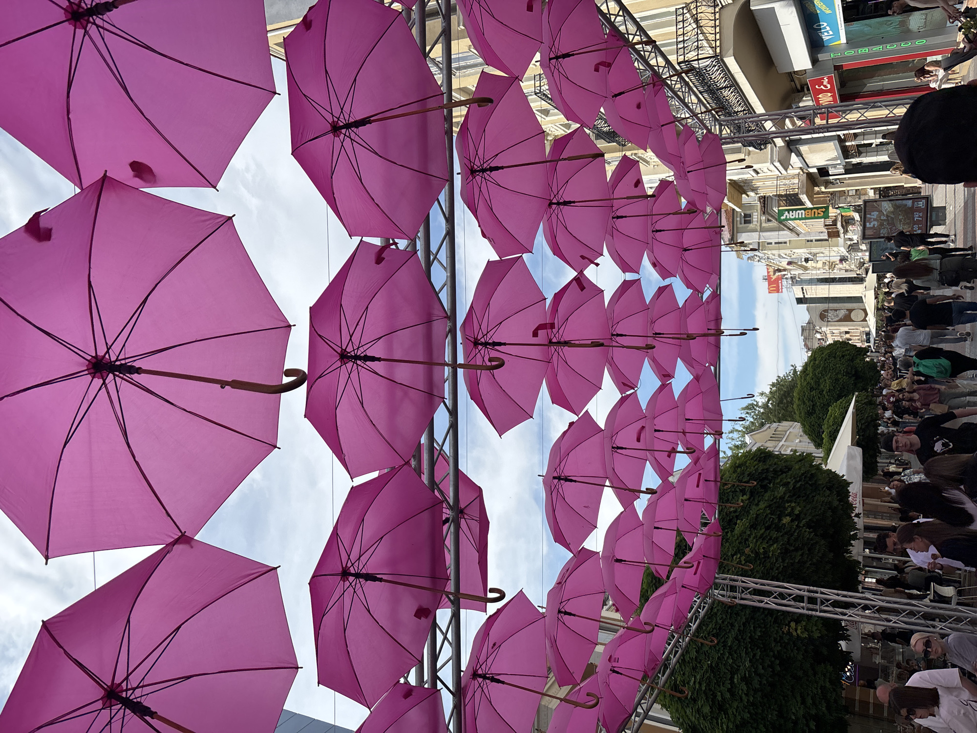 The image shows a street in the city of Unknown, Unknown, decorated with rows of pink umbrellas suspended overhead. These umbrellas are attached to a metal framework that spans the width of the street. The view is looking down the street, with buildings visible on both sides. A crowd of people is present, some walking along the street and others gathered in a seated area. Green trees and bushes are visible beyond the crowd. Signage for Subway and other businesses can be seen on the buildings. The sky is visible between the rows of umbrellas.