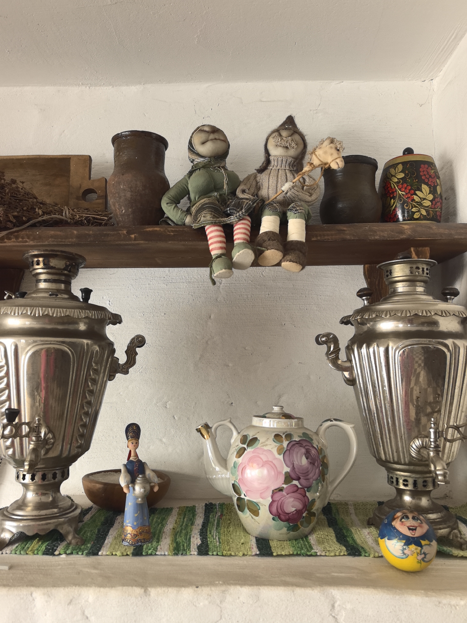 This indoor shot captures a shelf display filled with traditional Russian decorative items and kitchenware, hinting at a cozy, rustic atmosphere.
On the upper wooden shelf, from left to right, there's a pile of dried herbs or twigs next to a simple wooden cutting board. A tall, dark brown ceramic jug stands beside a fabric doll dressed in green with striped socks, sitting with its legs dangling. Next to it is another fabric doll, this one in beige with a mustache, holding a small stick horse. Further to the right, a smaller dark brown pot is visible, followed by a decorative wooden pot with a vibrant painted design featuring red berries and green leaves. The background wall is white and textured.
The lower shelf, also wooden, is covered by a green and white striped woven mat. Flanking this shelf are two large, shiny silver samovars, traditional Russian tea urns, each with a tap and ornate detailing. Between them, a small wooden figurine of a woman in a blue traditional dress and headdress, holding a miniature samovar, stands in front of a small wooden bowl. In the center, a white ceramic teapot is adorned with large, hand-painted pink and purple floral designs. To the right, near the second samovar, rests a small, yellow roly-poly doll with a cheerful, expressive face, often known as a Nevalyashka or Vanka-vstanka.
The scene depicts a static display, with no apparent activity. The items evoke a strong sense of Russian cultural heritage, fitting the provided location context of Chaikovsky. There is no visible text in the image.