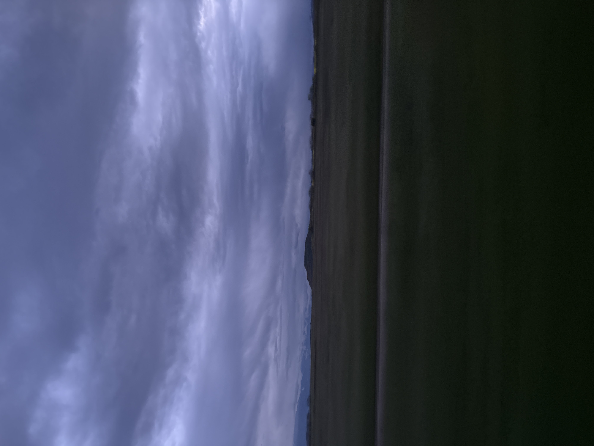 The image depicts a landscape in Tsar Asen, Bulgaria, under a cloudy sky. The left portion of the image showcases a dynamic, overcast sky filled with layers of gray clouds. To the right, the land is divided into two prominent sections: a darker, possibly waterlogged or shadowed area in the foreground and a strip of grassland running along the horizon. The grassland appears to be sparsely populated with trees. The horizon line is low and appears to separate the land from the sky. A few mountain peaks are discernible near the middle of the image.