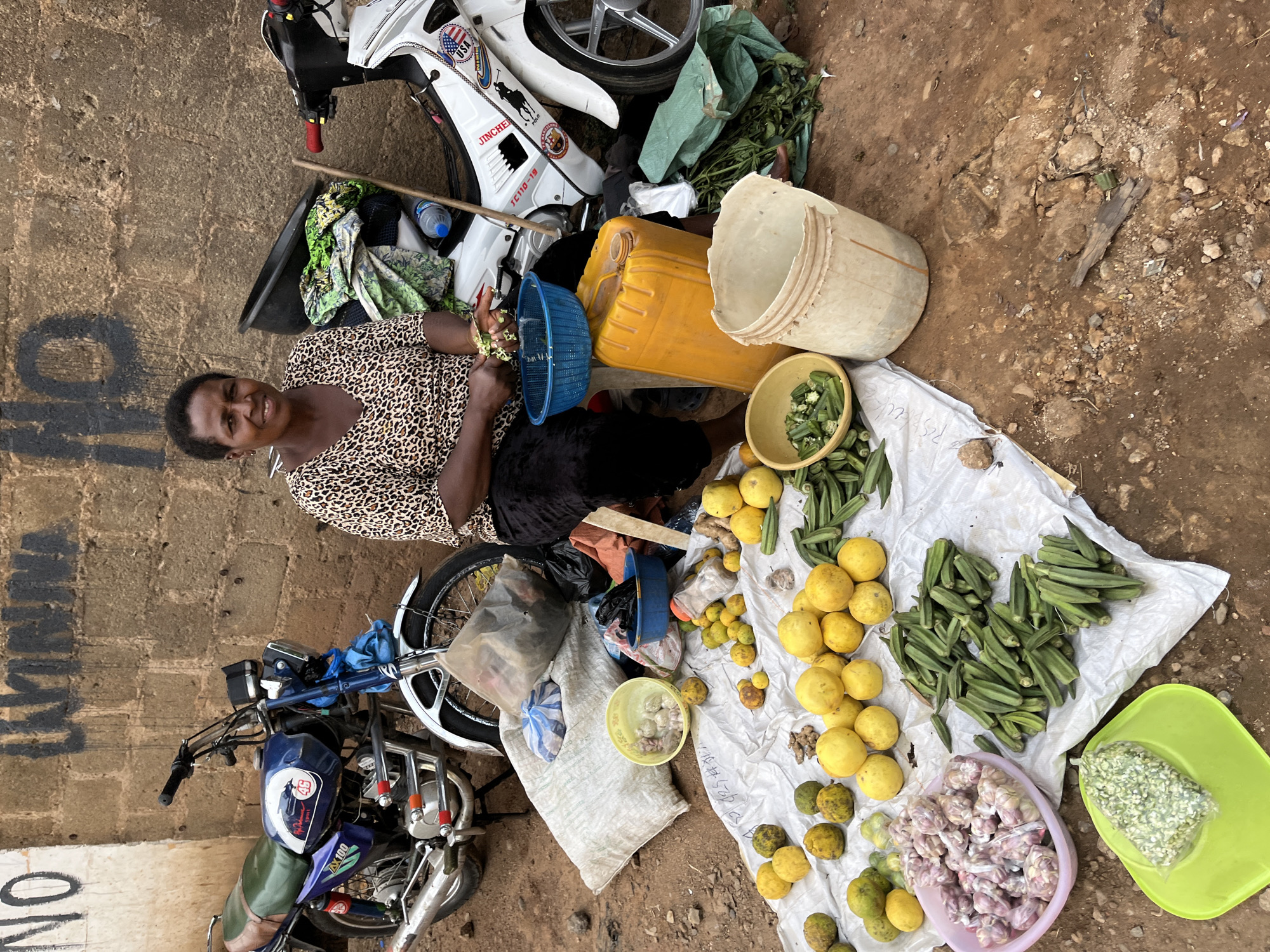 This image captures an outdoor market or roadside stall in Jos, Nigeria, showing a woman surrounded by various goods. The primary subject is a woman, likely a vendor, seated on the ground in the upper-left quadrant of the frame. She wears a leopard-print top and dark attire, looking directly at the viewer with a friendly smile. Her hands are near a blue plastic colander, suggesting she is sorting or preparing produce.
Spread out on a white sheet on the dirt ground before her is an assortment of fresh produce. This includes numerous yellow-skinned fruits resembling lemons or limes, clusters of green okra pods, ginger roots, and other green, round fruits. Adjacent to these are a light purple bowl filled with small, light-colored bulbs (possibly garlic), and a bright lime-green plastic tray holding a clear plastic bag containing finely chopped green vegetables.
Around the woman and her display are several utilitarian objects: a large yellow jerrycan, a tall white plastic bucket, and a smaller yellow bowl with cut okra. Two motorcycles are visible: a white one parked behind her, bearing "USA," "JINCHEN," and "JC110-19" text along with a Polo logo, and a blue one positioned towards the lower left, displaying "AX100" and the number "46." The ground is uneven dirt, and a wall runs along the left edge of the scene, marked with partially visible black graffiti that appears to be "14" or "91" (when rotated) and "NO." The scene suggests a busy daytime environment, typical of an informal market setting.