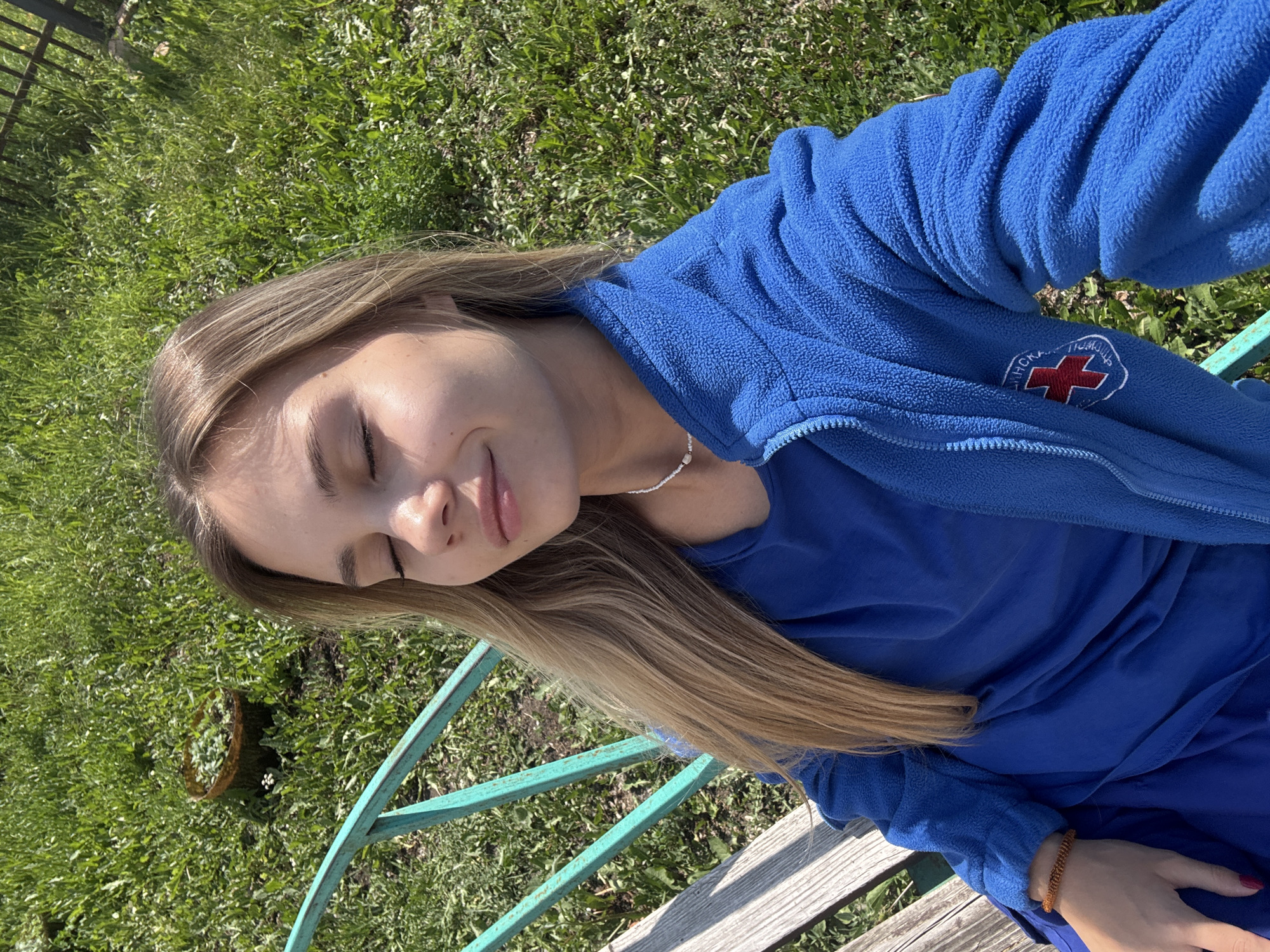 The image is a selfie of a young woman with long blonde hair, wearing a blue fleece jacket with a red and white emblem on the left sleeve. She is sitting on a wooden bench with a green metal frame, outdoors. Her eyes are closed, and she has a serene expression on her face. The sun is shining, casting light on her skin and hair, suggesting it is daytime. The background is a grassy area with some greenery and a small plant in a pot visible to the left. A fence is partially visible in the upper left corner of the image. The emblem on her jacket appears to have Cyrillic text, but it is not fully legible. The setting and the presence of a red cross emblem on the jacket might suggest a connection to a medical or volunteer organization, possibly in Chaikovsky, Russia, given the location context. The overall mood of the image is peaceful and relaxed, with the woman enjoying the sunny weather.