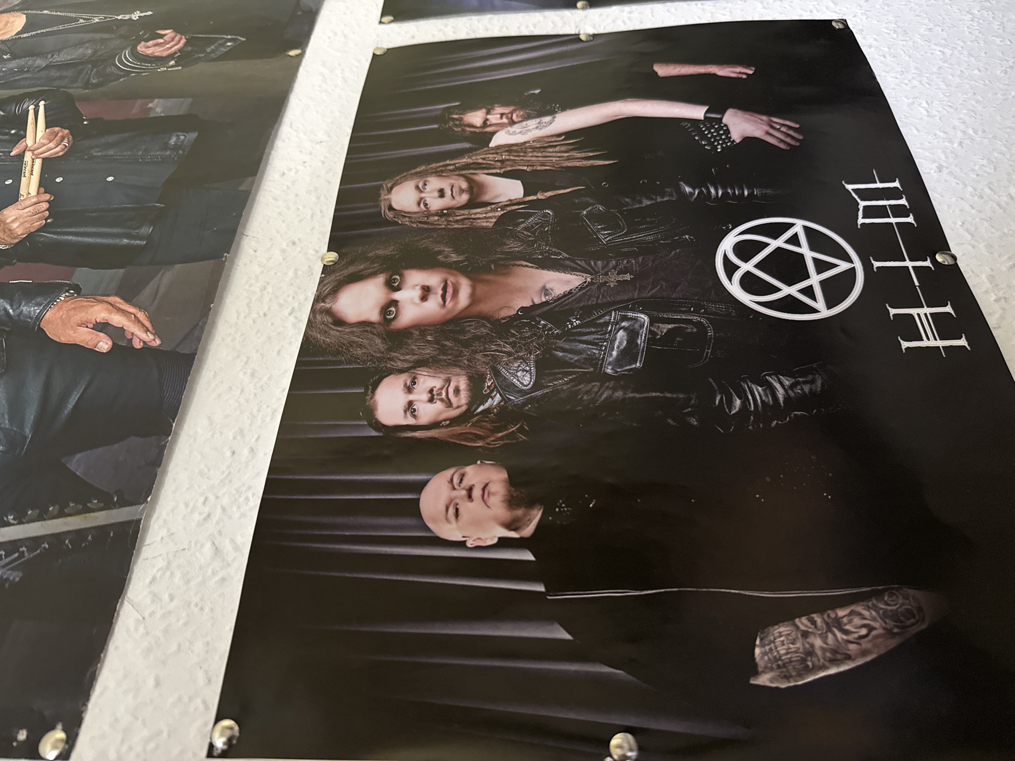 The image shows a poster of the band HIM hanging on a white, textured wall, held up by visible push pins. The poster depicts five male band members in black attire against a dark curtain backdrop. The band members are identified by the HIM logo and their stylized band name in white text at the bottom of the poster. The band members are positioned from left to right, with the leftmost member having a shaved head and beard, and visible tattoos on his arm. The central figure has long, dark, curly hair and is wearing a cross necklace. The member to his right has long, braided hair. The other two members are partially visible. The poster is mounted at eye level in what appears to be a domestic or casual setting, likely a bedroom or living area. Based on the provided information, this poster is located in Bad Sachsa, Germany.