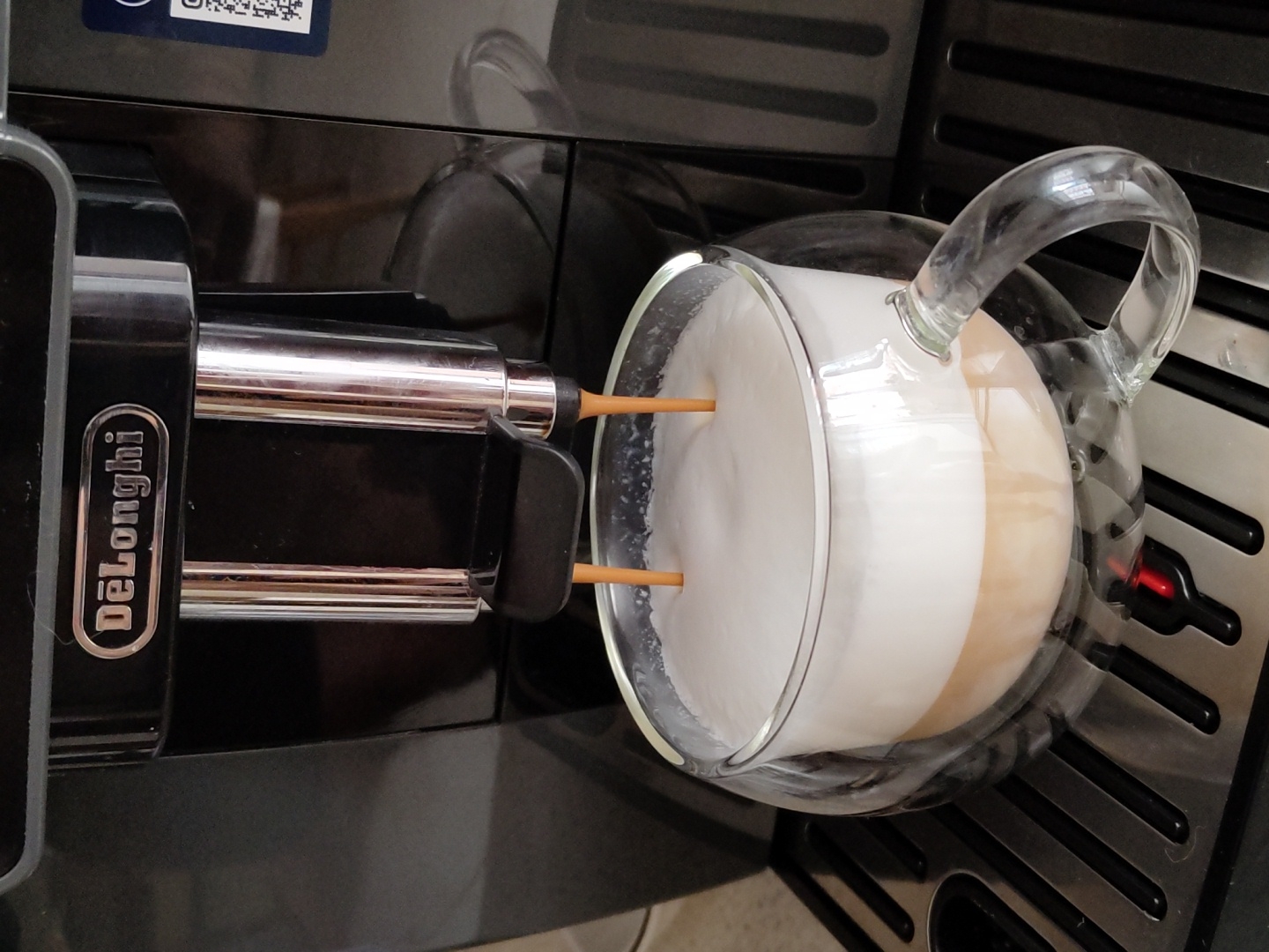 A close-up, eye-level shot captures a De'Longhi coffee machine dispensing a latte into a clear glass mug. The coffee machine is black with chrome accents, and the De'Longhi logo is visible on the front. Two streams of liquid, one dark brown and one white, are pouring from the machine's spouts into the mug, which is already filled with layered coffee and foam. The mug has a clear handle and sits on the machine's drip tray, which is black with silver slats and a red indicator light. The background is dark and out of focus, suggesting an indoor setting. There are no people or animals visible in the image. The image is crisp and well-lit, focusing on the coffee-making process. The scene is consistent with a home or small cafe environment, and no specific location within Krasnodar, Russia, can be identified.