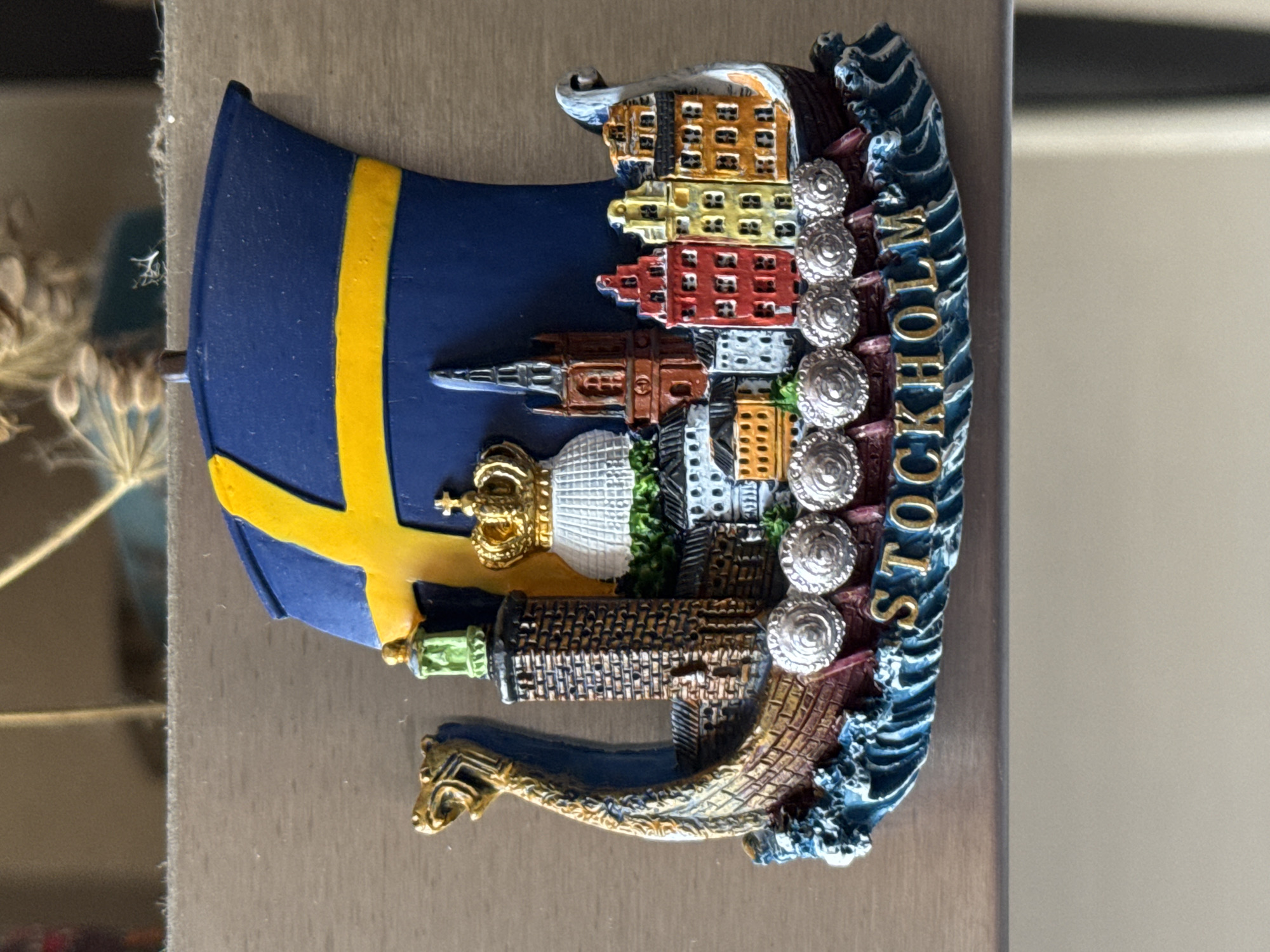 A souvenir magnet in the shape of a Viking ship is displayed on a light-colored, textured surface, likely a refrigerator. The ship features a blue sail with a yellow cross, representing the Swedish flag. Adorned on the deck are miniature representations of Stockholm's landmarks, including a tall brick tower with a green lantern, a white domed building topped with a golden crown, and several colorful buildings with pointed roofs. The ship's prow is carved in the likeness of a dragon's head. The words "STOCKHOLM" are inscribed in gold letters along the base of the magnet, which is shaped to resemble choppy blue waves. The overall scene is well-lit, with the magnet being the sole focus against a neutral background. The location is not Bishkek City, Kyrgyzstan, as there are no identifiable elements of that city present.